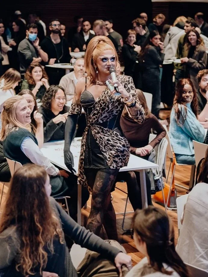 Une drag queen avec des vêtements à motif léopard, portant des gants noirs et tenant un microphone, entourée d'un public assis et debout dans une salle.