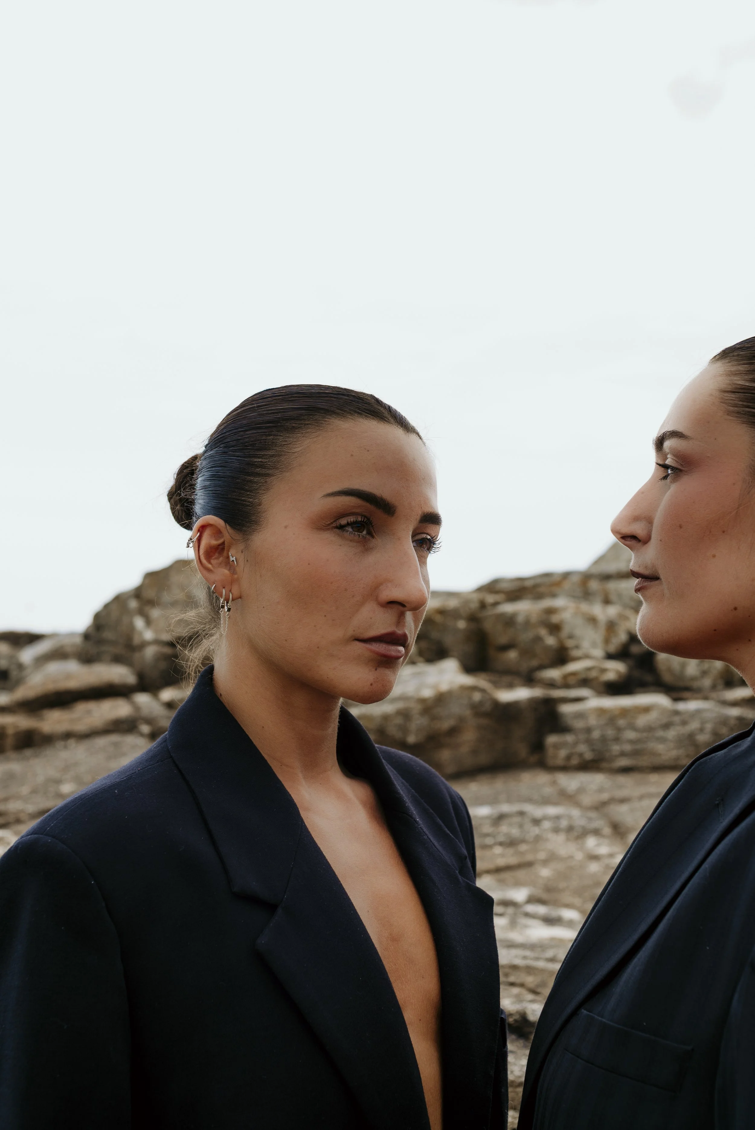 Two women standing face to face outdoors with rocky terrain in the background, both wearing dark blazers, with their hair slicked back, gazing intently at each other.