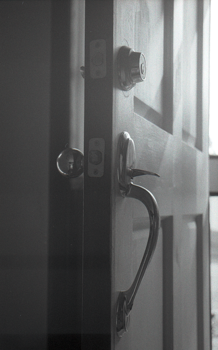 A black and white photo of a partially opened door with a shiny metal handle, door lock, and deadbolt, with sunlight coming through the window in the background.