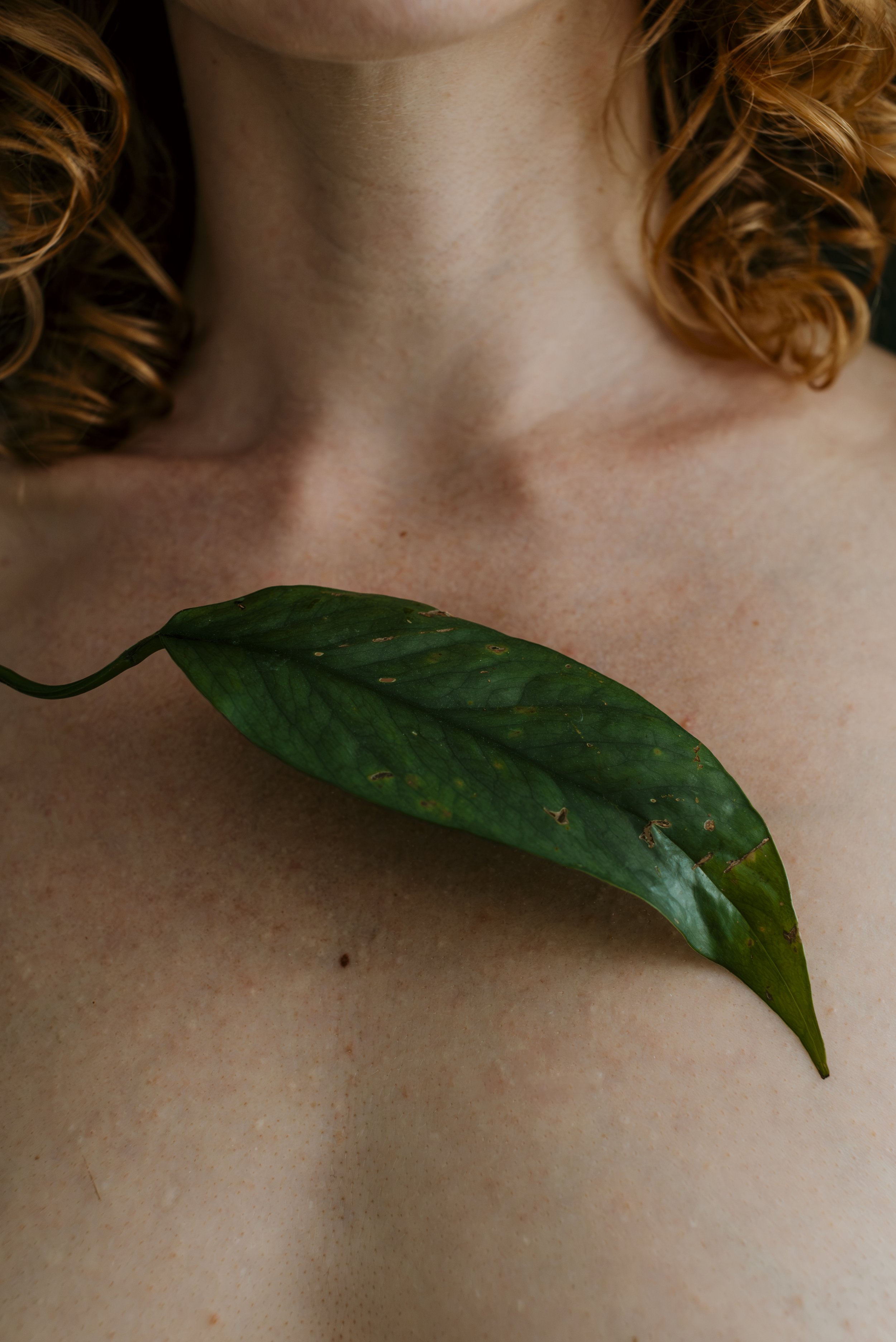 Close-up of a person's upper chest and neck with red, curly hair visible on the sides. A large green leaf is placed horizontally across the chest, partially covering the person's skin.