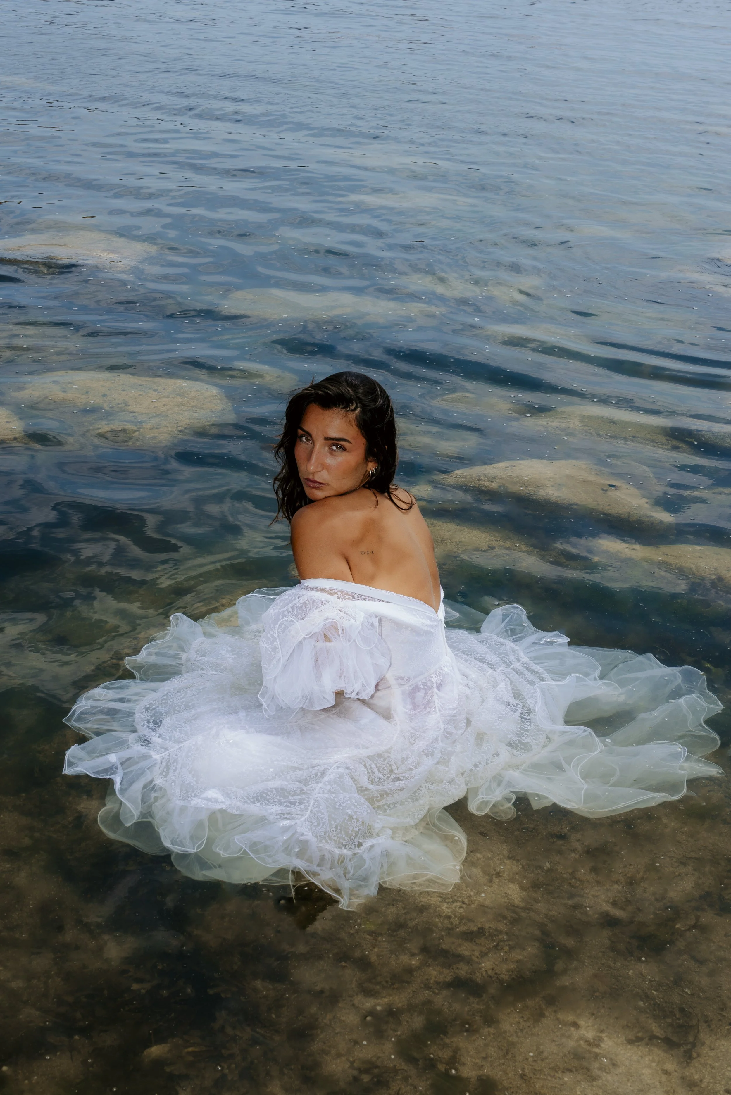 A woman sitting in clear, shallow ocean water, wearing a white dress with ruffled fabric, looking at the camera with her long dark hair loose.