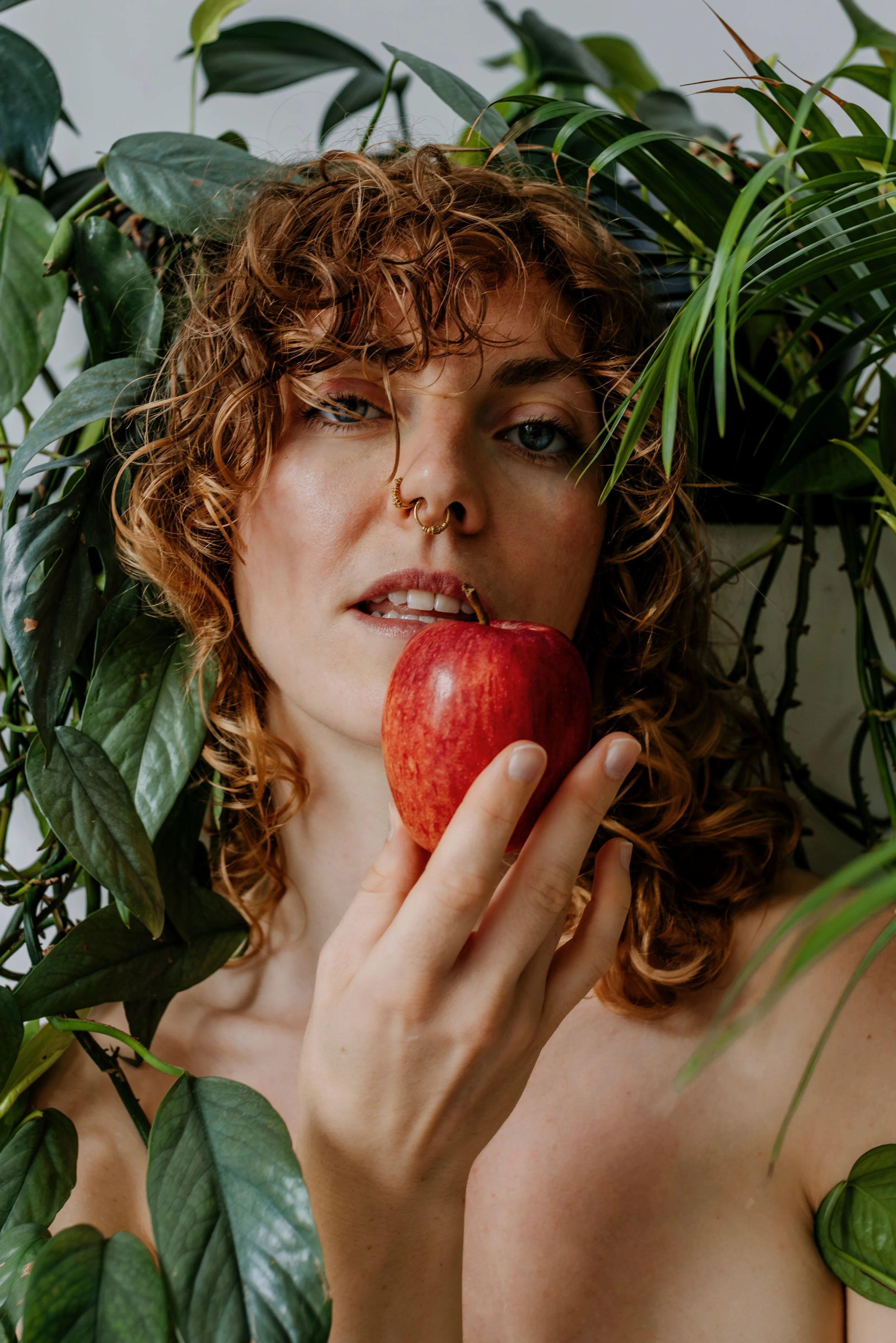 A woman with curly red hair and blue eyes holds a red apple near her mouth, surrounded by green leafy plants.