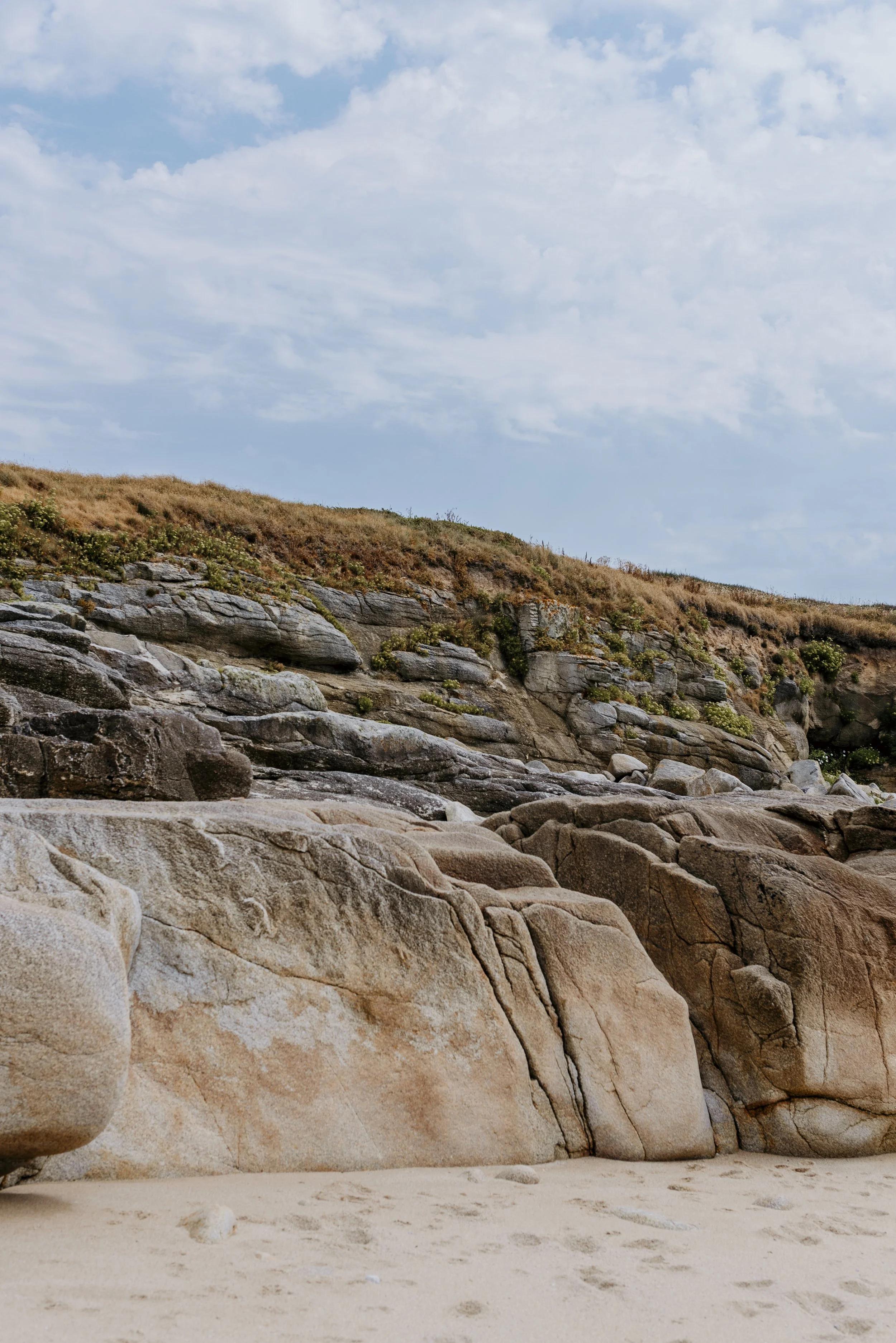 A rocky coastal landscape with large, smooth rocks and a sandy beach in the foreground. There is a hillside with sparse vegetation and a partly cloudy sky in the background.