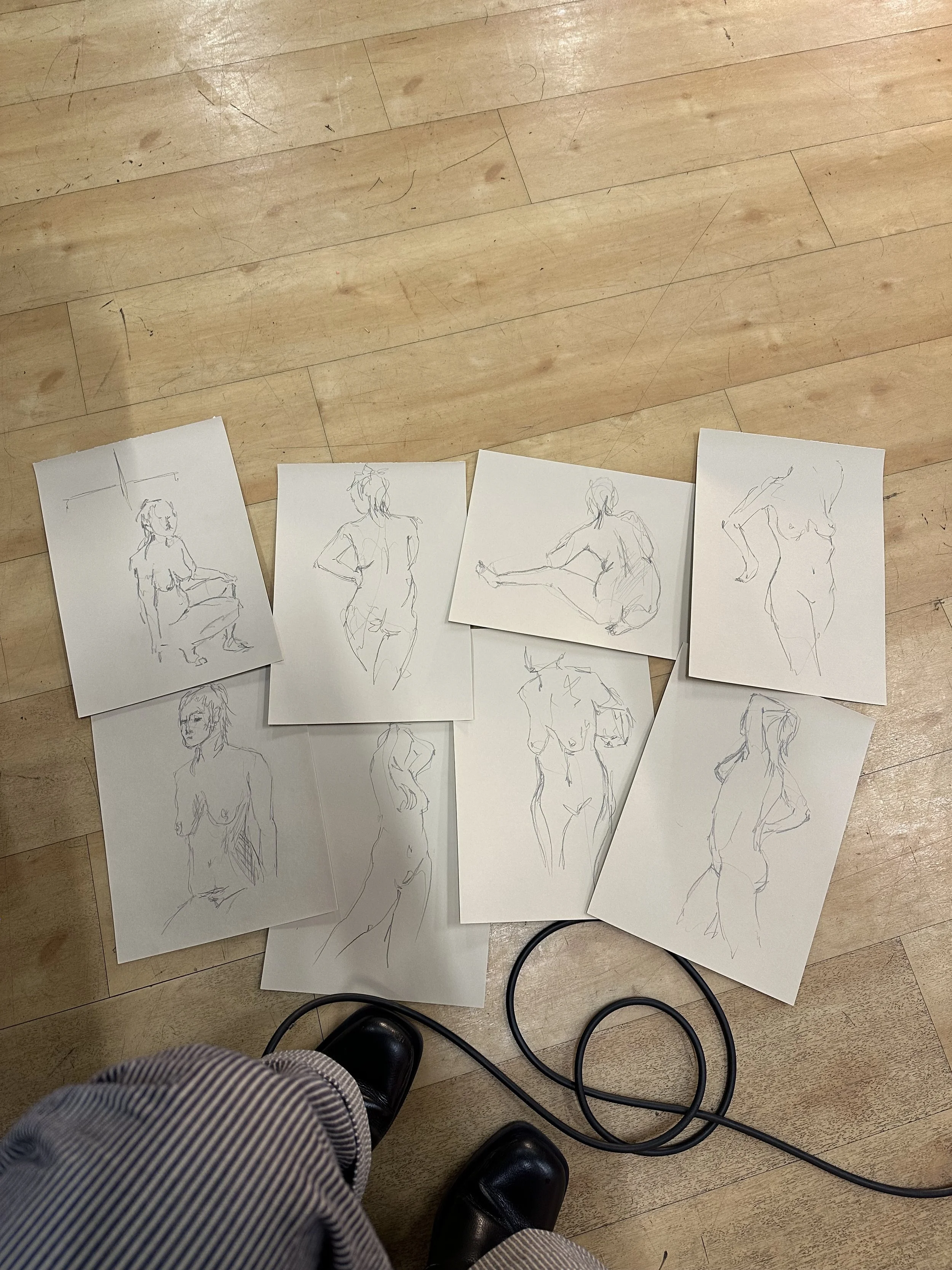 Nine quick sketches of female figures on paper, spread on wooden floor. The sketched figures are in different poses, some sitting, standing, and leaning, with a pair of black shoes and a black electrical cord visible at the bottom.