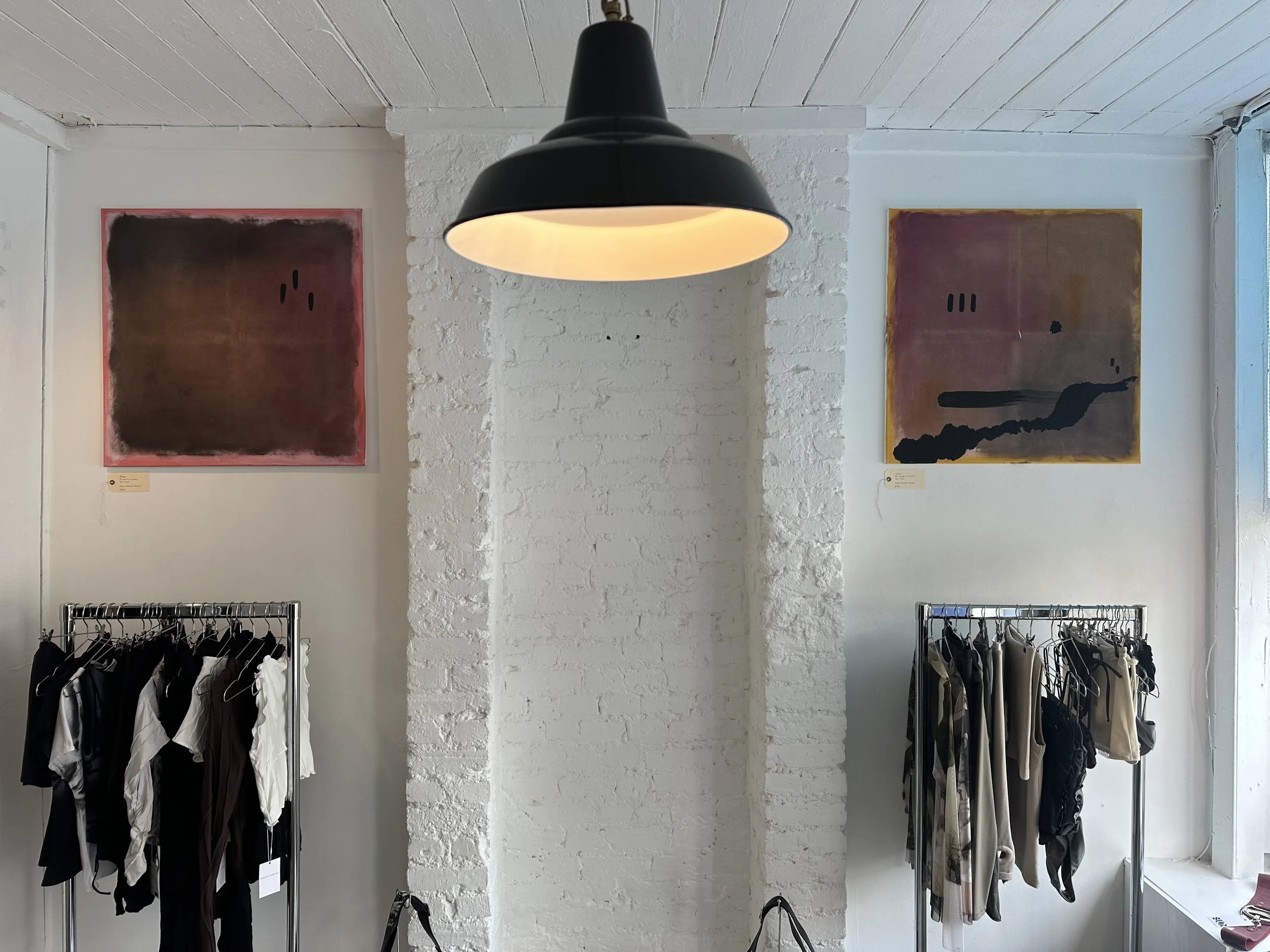 A retail clothing store interior with two clothing racks filled with various garments, a black pendant light hanging from the ceiling, and two abstract paintings on the white brick wall behind the racks. Sunlight streams in from a window on the right.