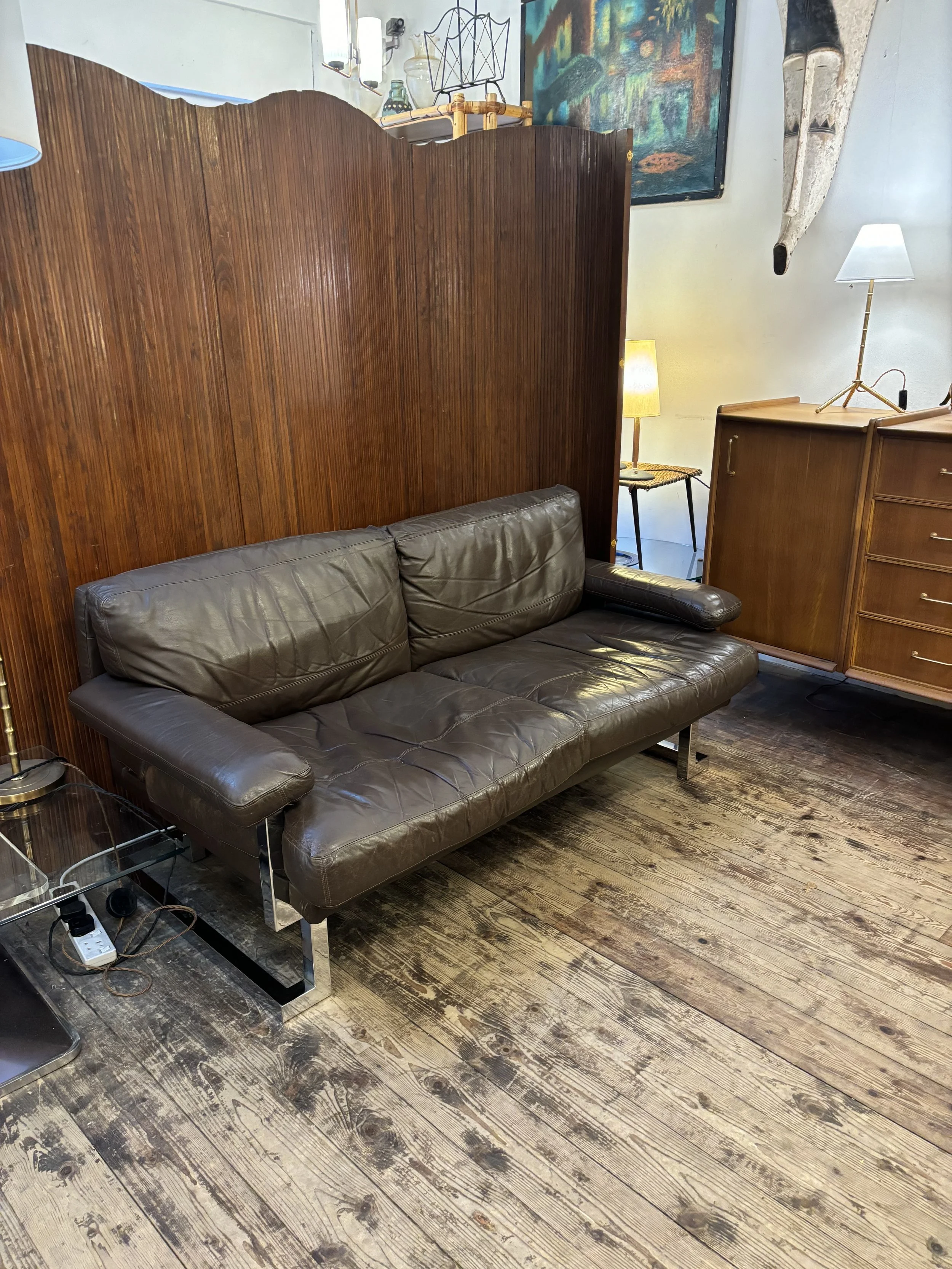 A vintage brown leather sofa with metal legs in front of a wooden room divider. To the right, a wooden dresser with a white lamp on top, placed on a wooden floor. In the background, there's a lamp with a yellow shade, a chair, and wall art with a framed painting and what appears to be a rolled-up rug hanging on the wall.