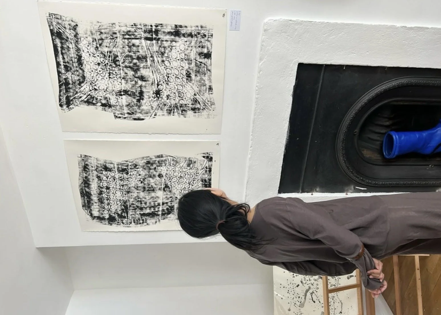 A woman with dark hair tied back, wearing a gray coat, stands with her hands clasped behind her back, observing black-and-white abstract artworks displayed on a white wall.