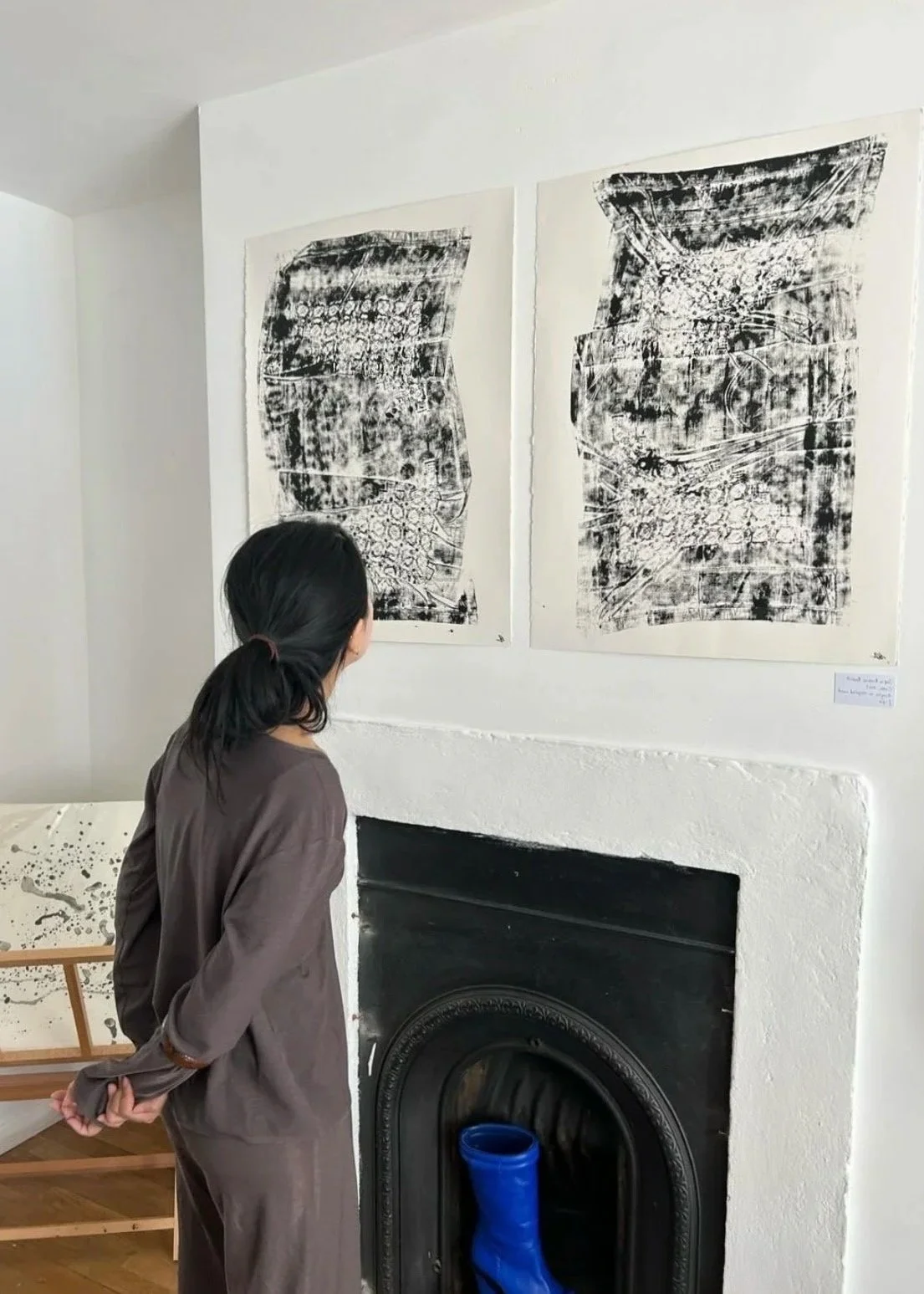 A woman with black hair tied back, wearing a brown dress, stands in an art gallery looking at two abstract black and white paintings hung on a white wall above a black fireplace with a blue object inside.