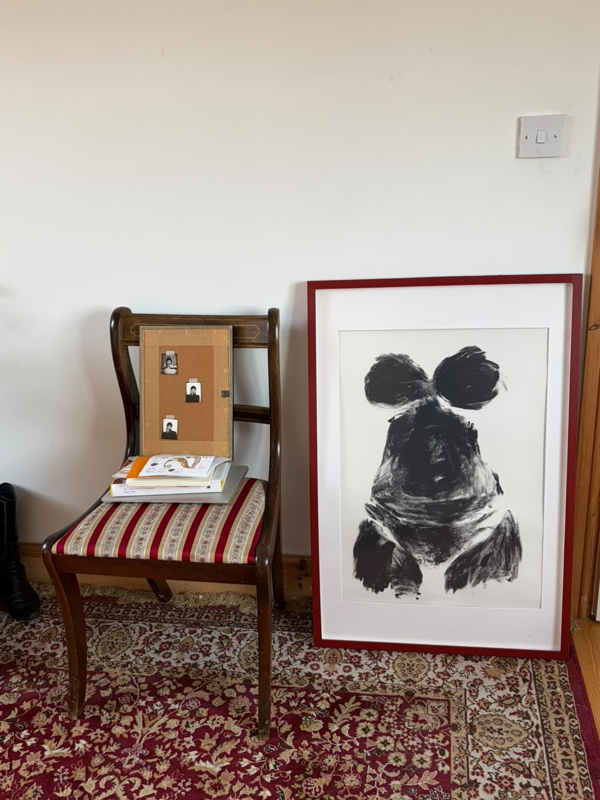 A wooden chair with a cushioned seat, a framed painting of a black abstract dog brushstroke leaning against the wall, and a small corkboard with four black and white photographs pinned to it, all on a patterned area rug.