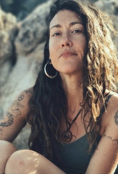 A woman with long, wavy hair and tattoos, wearing a sleeveless top, sitting outdoors with rocks in the background.