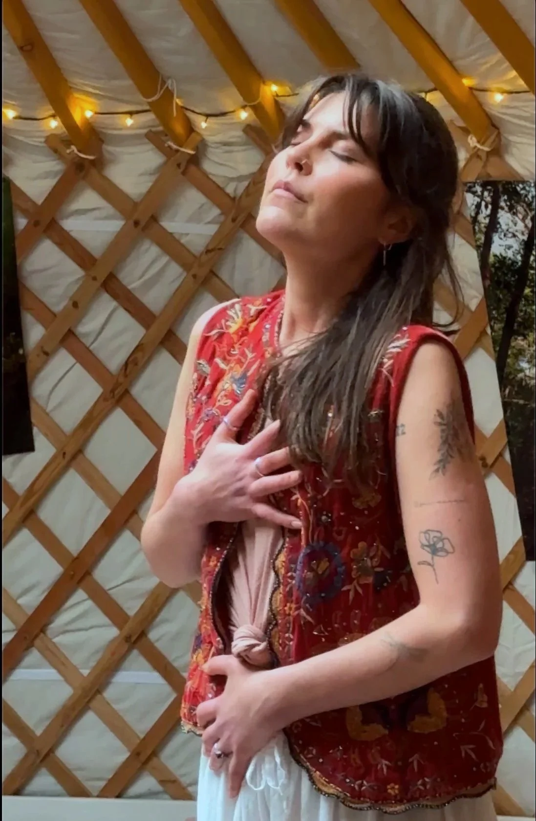 A woman with long brown hair and tattoos on her arms, wearing a red embroidered vest and white pants, with her eyes closed and one hand on her chest, in front of a wooden lattice with string lights.