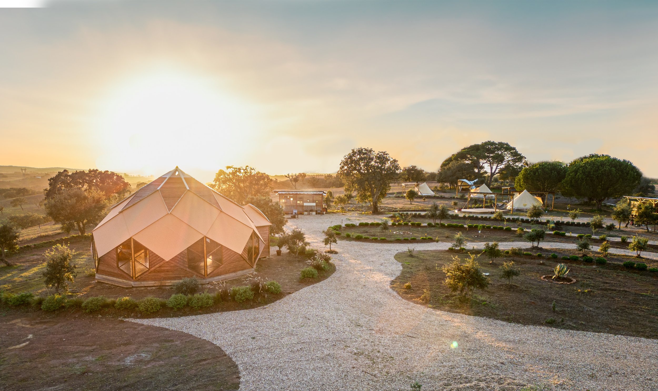 A landscape at sunset featuring a modern geodesic dome structure, gravel pathways, scattered trees, and small tents or canopies in a serene outdoor setting.