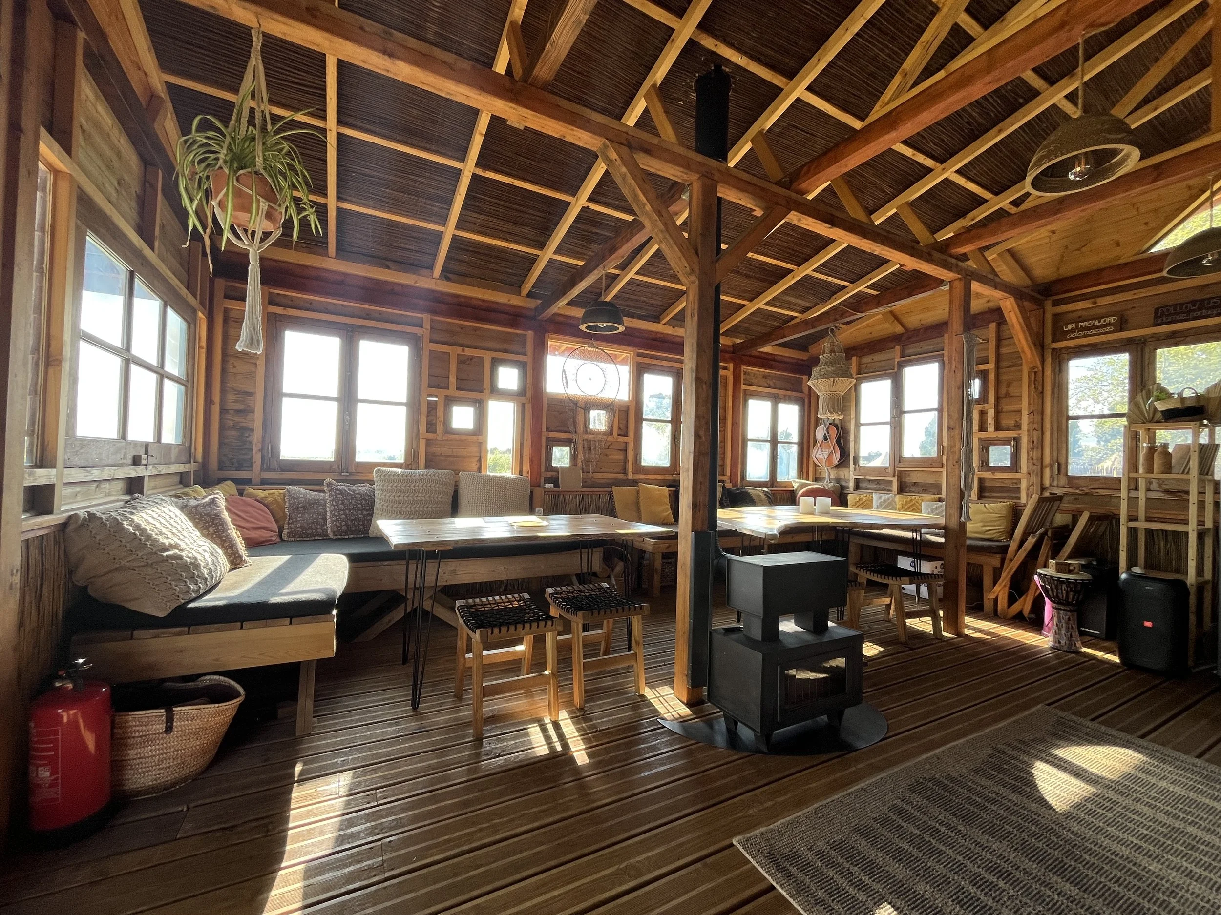 Interior of a cozy wooden cabin with large windows, built-in benches with cushions, a wood stove, and hanging plants. Sunlight illuminates the space, creating a warm and inviting atmosphere.