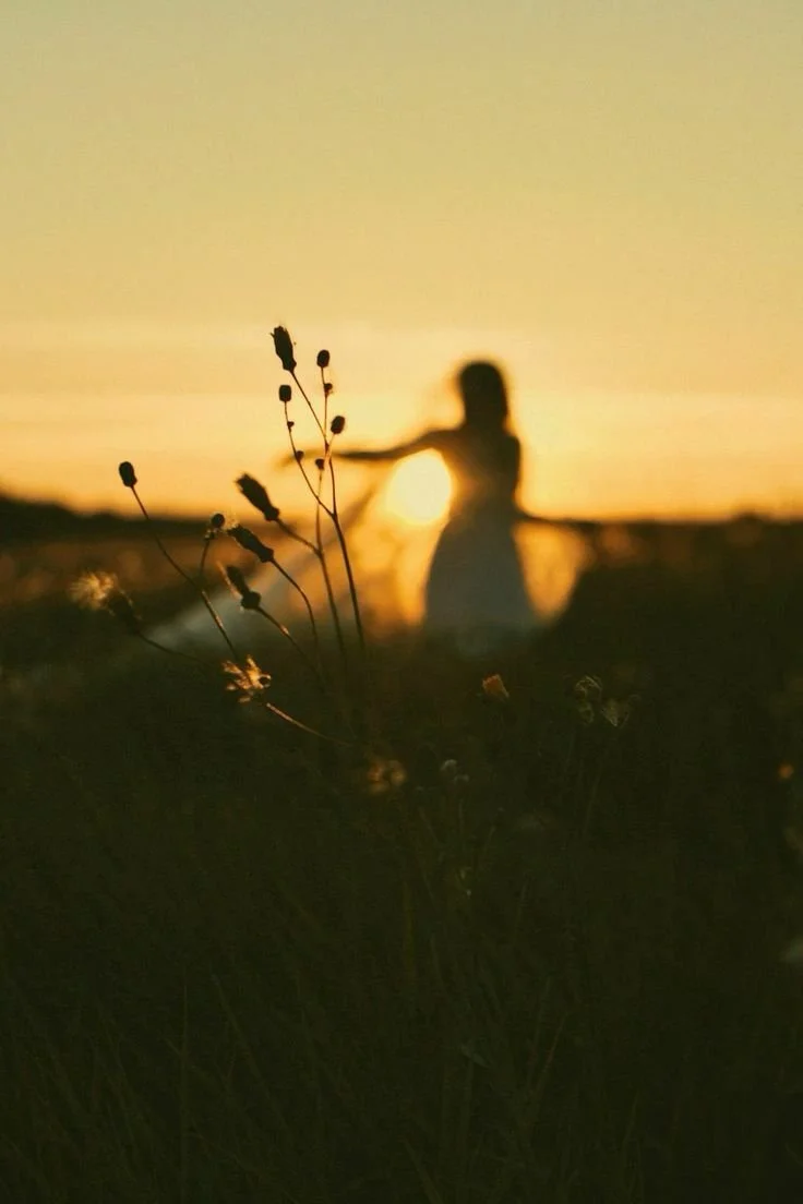Silhouette of a woman extending her arms during a sunset, with grasses in the foreground and a clear sky in the background.