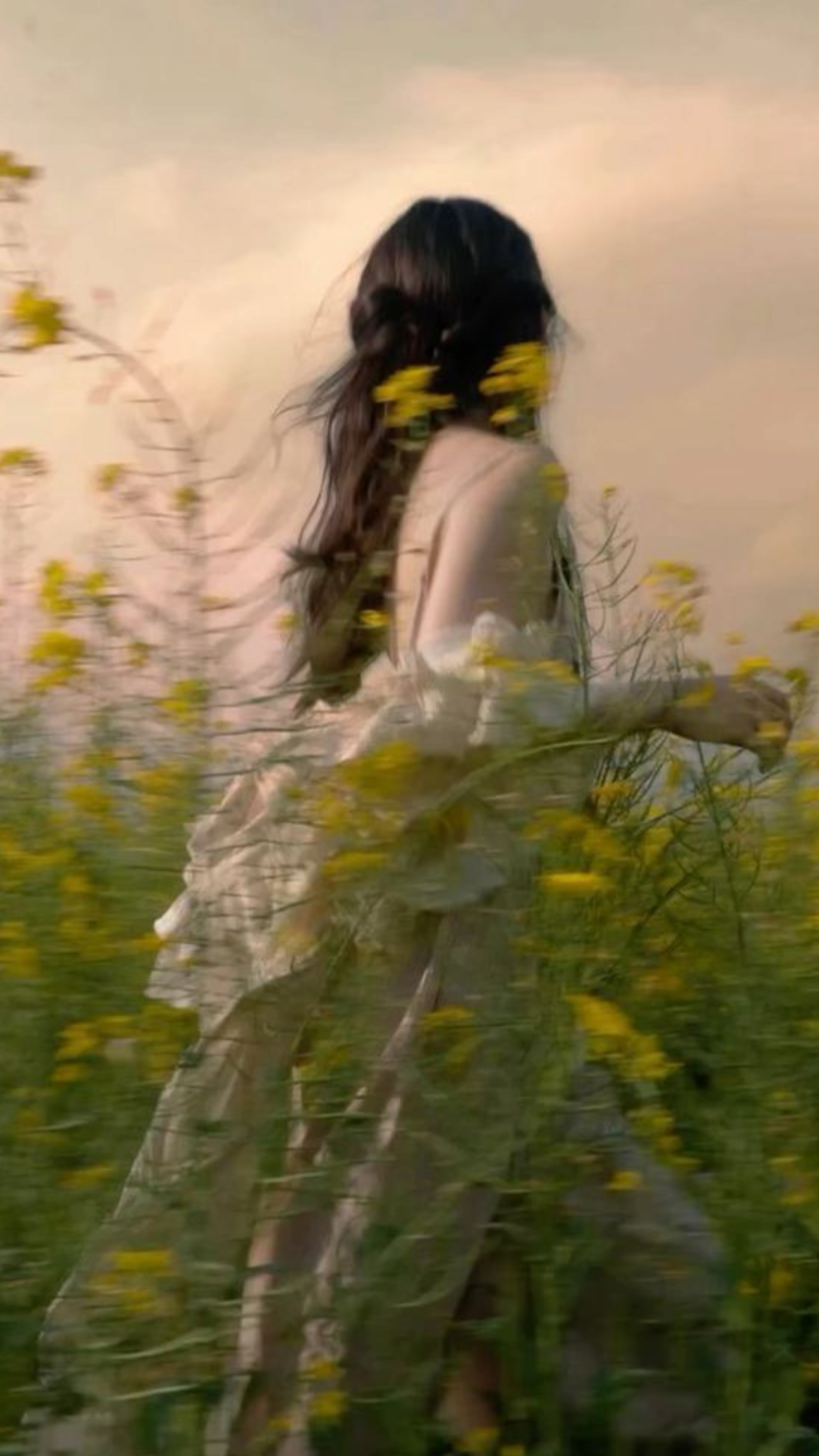 Blurry photo of a woman with long dark hair walking or standing among yellow wildflowers, facing away from the camera, with a subdued sky background.