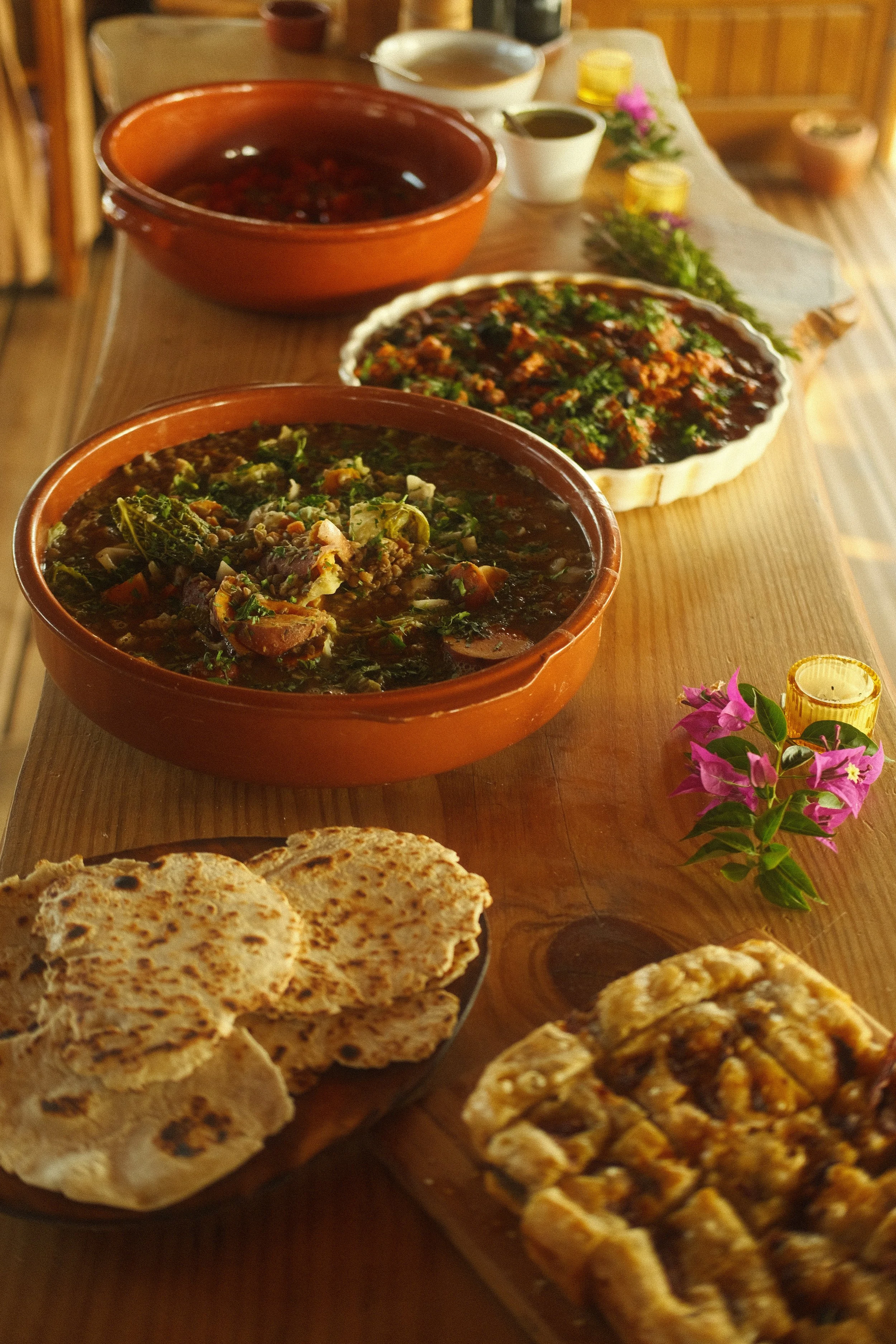 A traditional Indian meal spread on a wooden table, including two bowls of vegetable curry, naan bread, and a pie-like dish, with small bowls of sauces, a flower, and a candle for decoration.