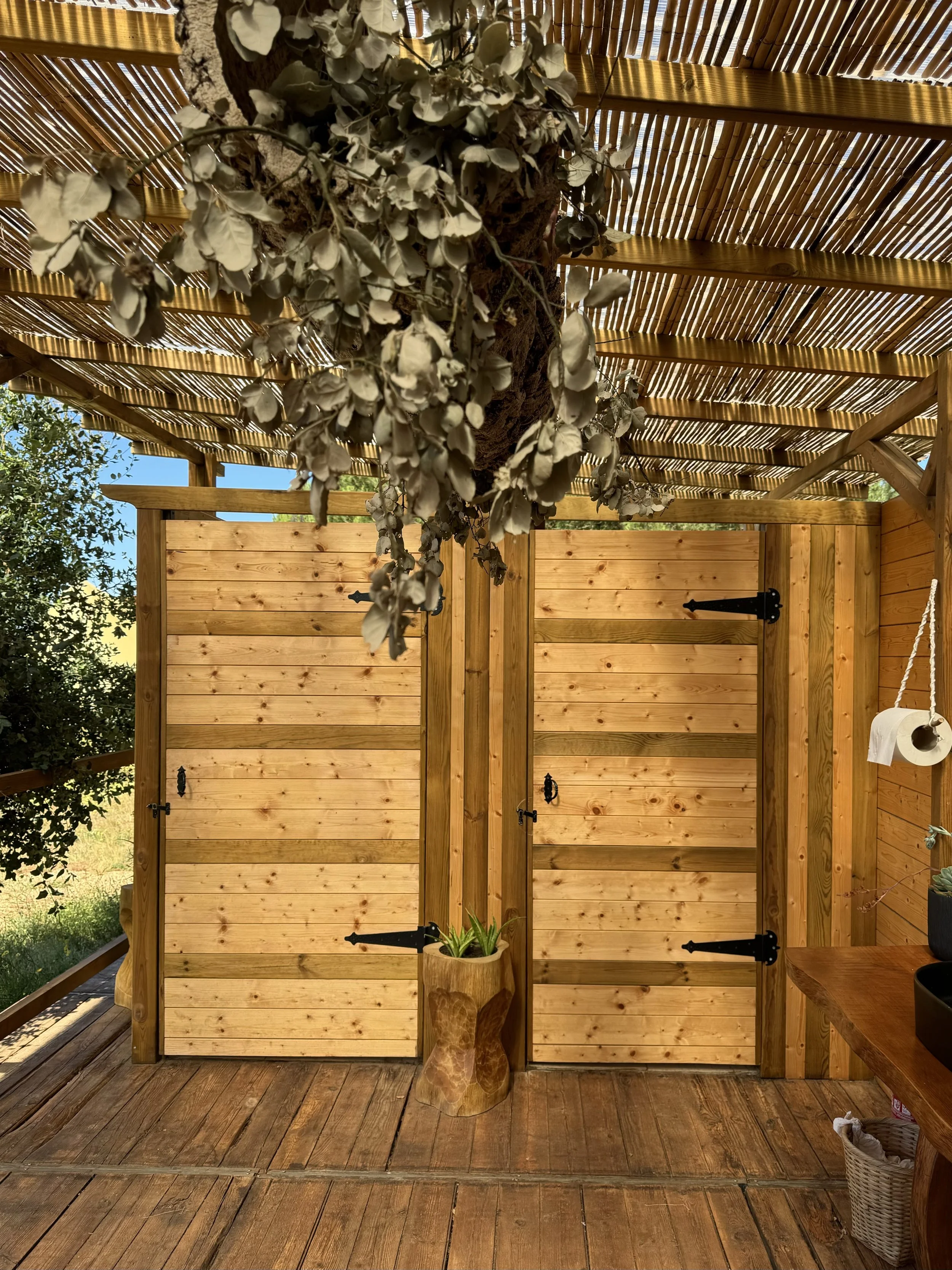 Wooden outdoor shower with two closed doors, hanging black handles, under a bamboo roof, with a hanging toilet paper roll and potted plants on a wooden counter.