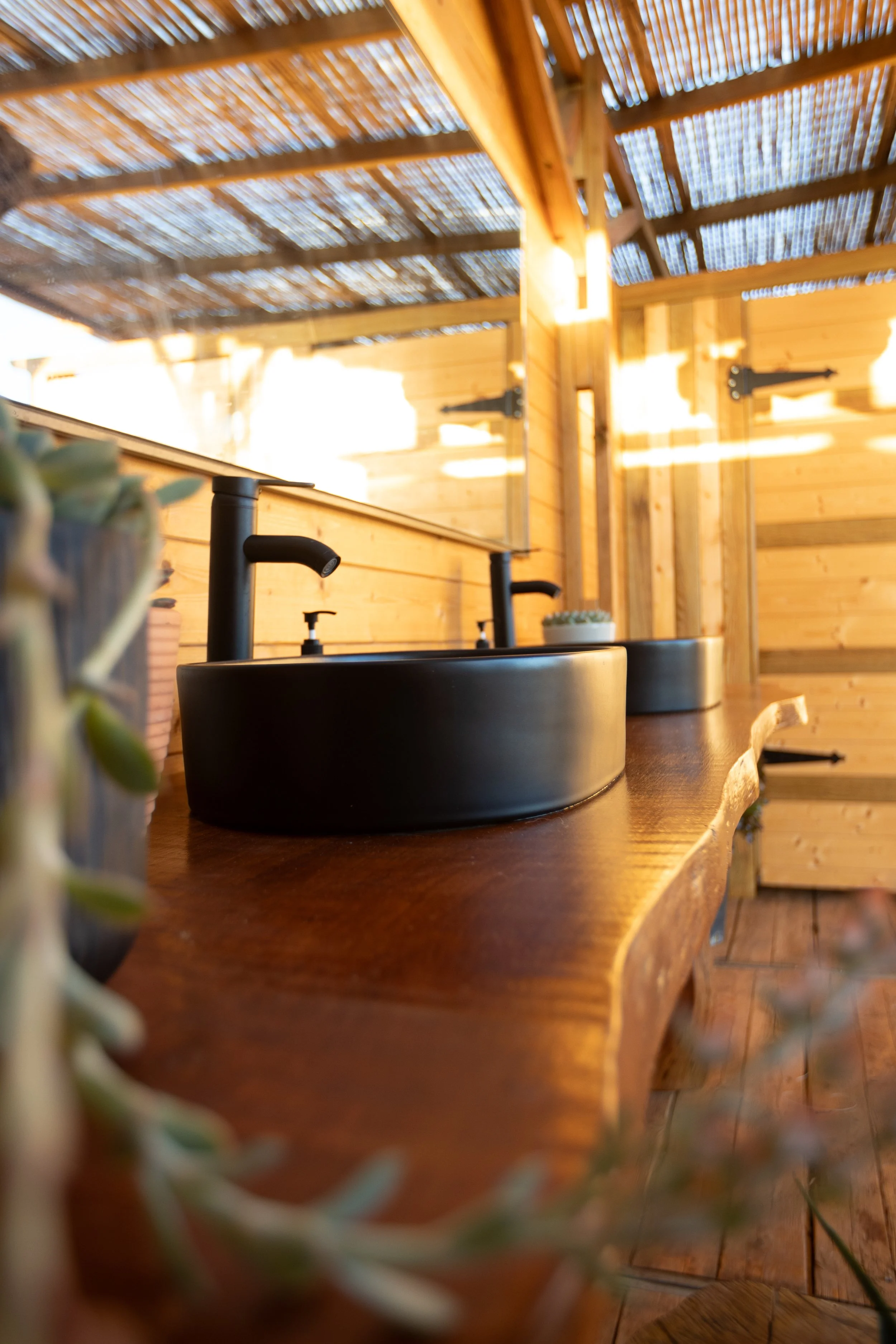 A rustic wooden bathroom vanity with two black vessel sinks and black faucets, set against wooden walls and a bamboo ceiling in a cozy outdoor or semi-outdoor setting.