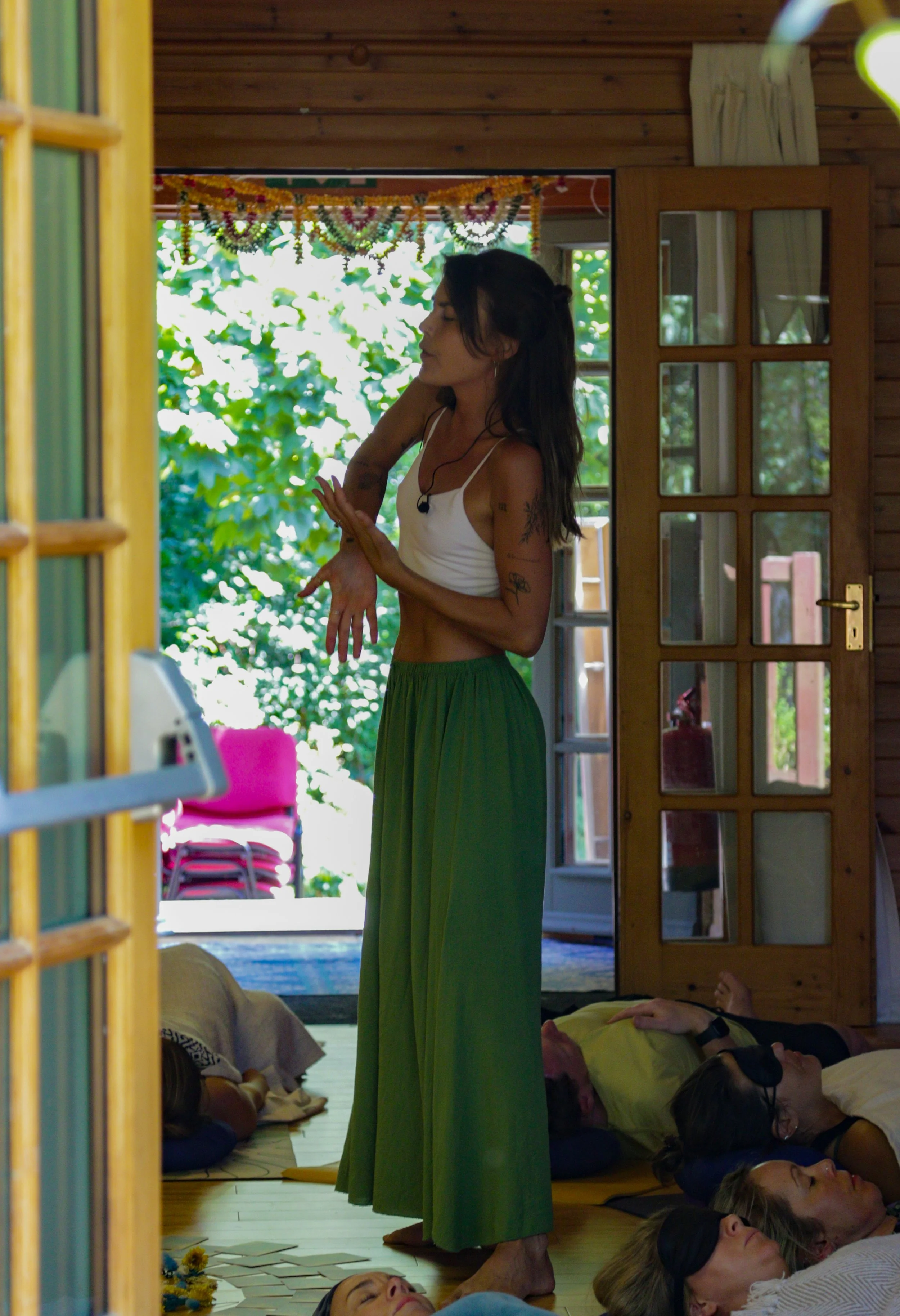 A woman standing in a room, leading a meditation session with people lying down on the floor. The room has wooden walls and an open door revealing greenery outside.