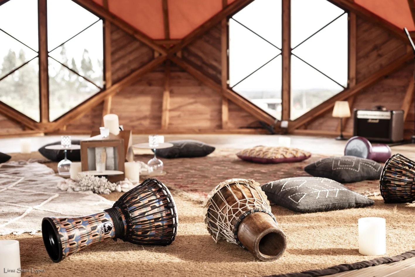 Interior of a cozy wooden room decorated with cushions, candles, drums, and a speaker, with large windows letting in natural light.
