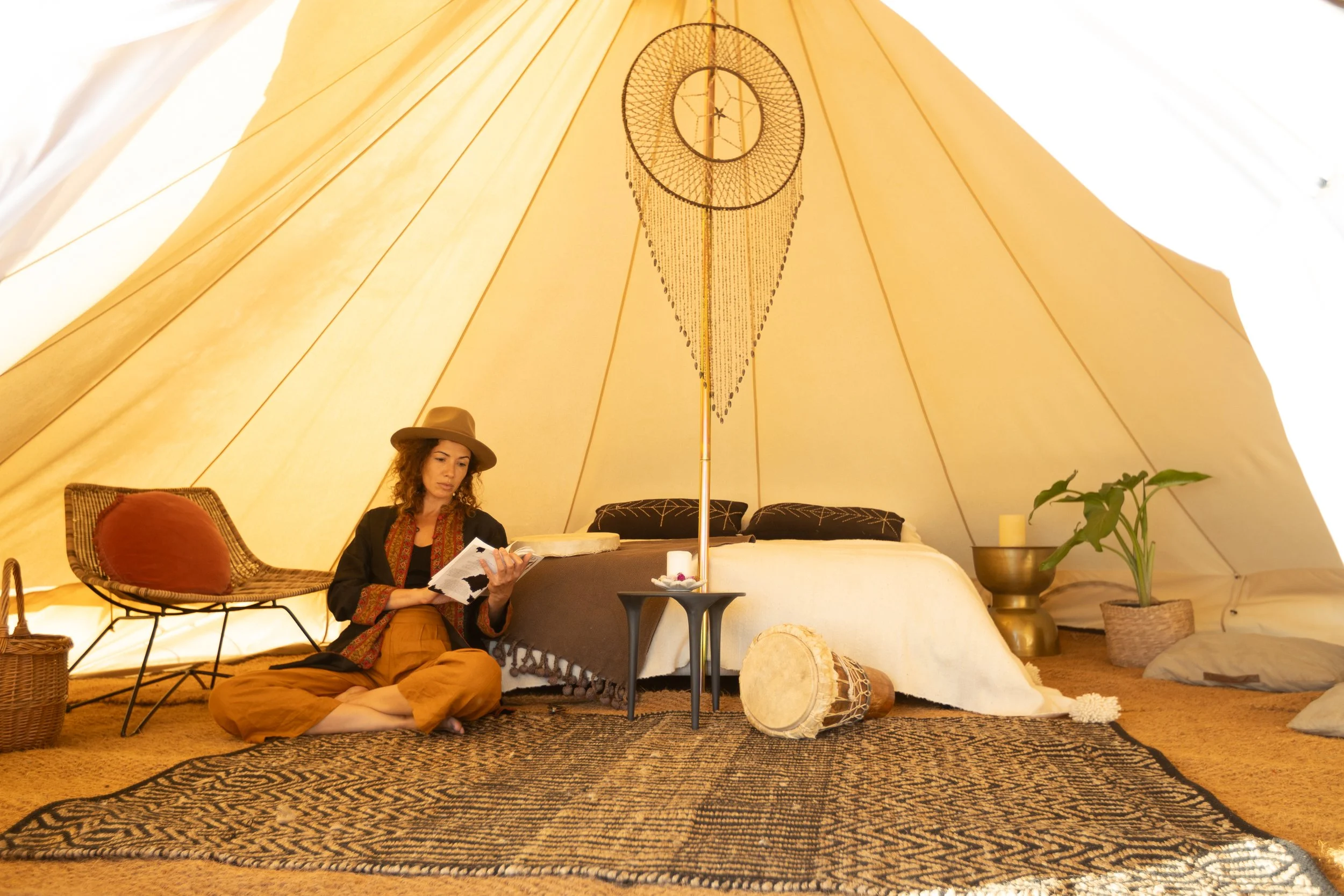 A woman sitting cross-legged inside a beige camping tent, reading a book. The tent is decorated with a dreamcatcher hanging from the pole, and has a bed with pillows and blankets. There is a woven chair with a pillow, a small side table, a potted plant, a drum, and a patterned rug on the floor.