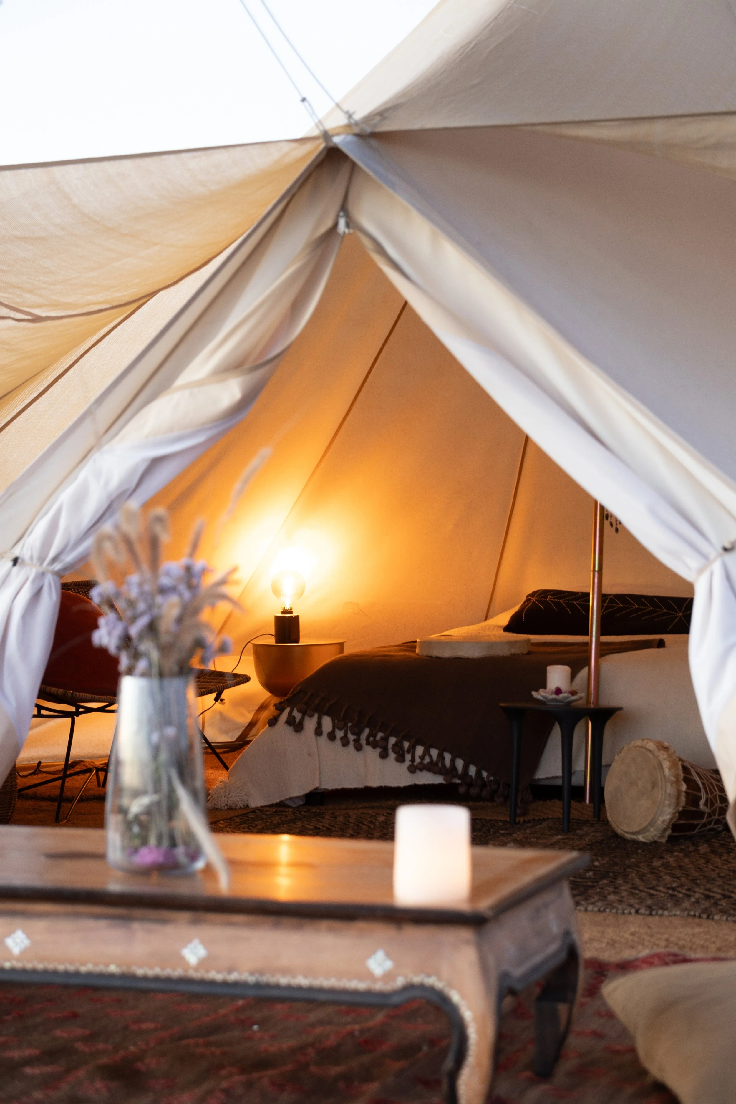 Cozy tent interior with a bed, lit by a warm lamp, and decorative items like a candle and flowers on a wooden table.