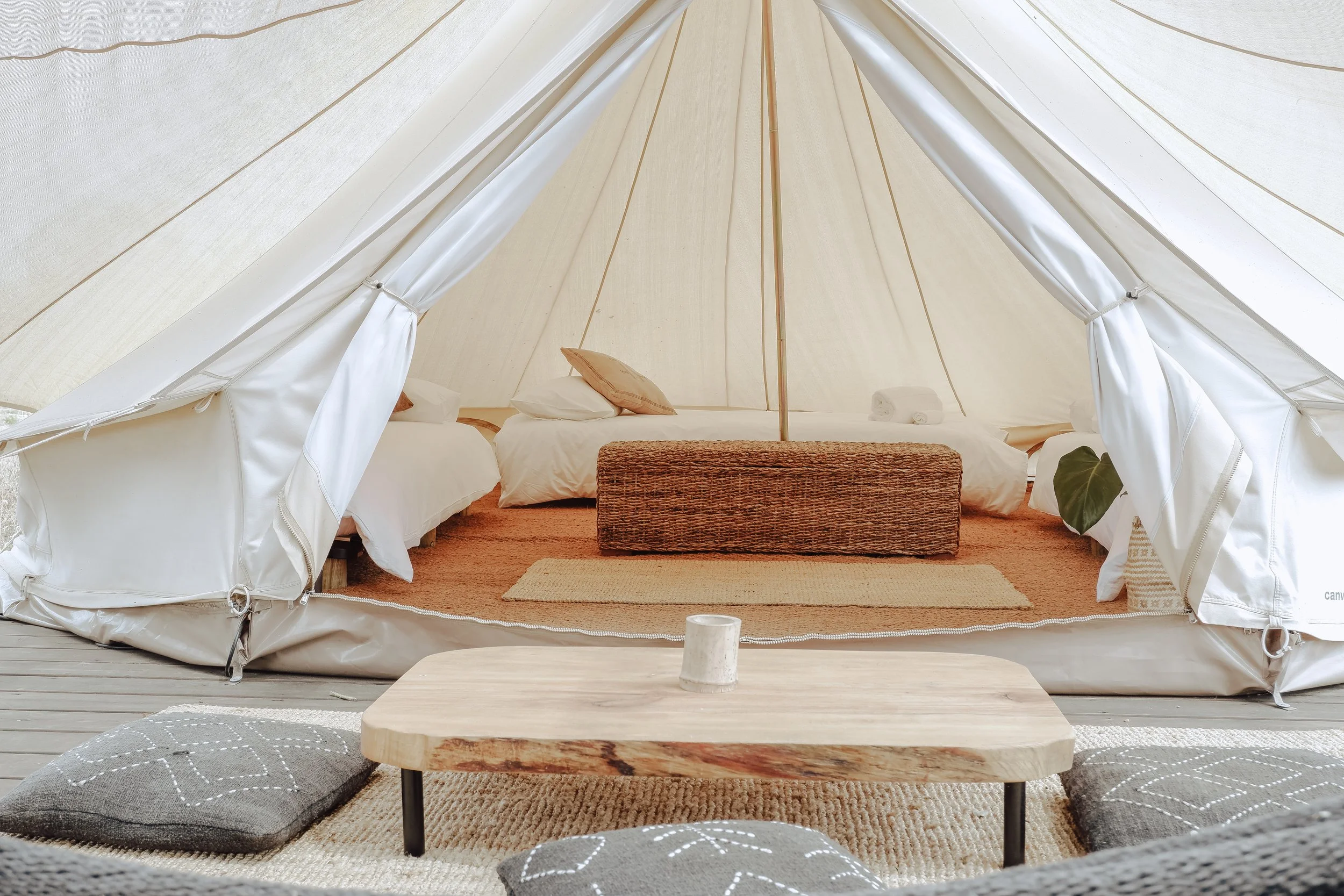 Interior of a beige canvas tent with a bed, pillows, and rolled towels, seen from the front with a low wooden table and cushions on a rug outside the tent.