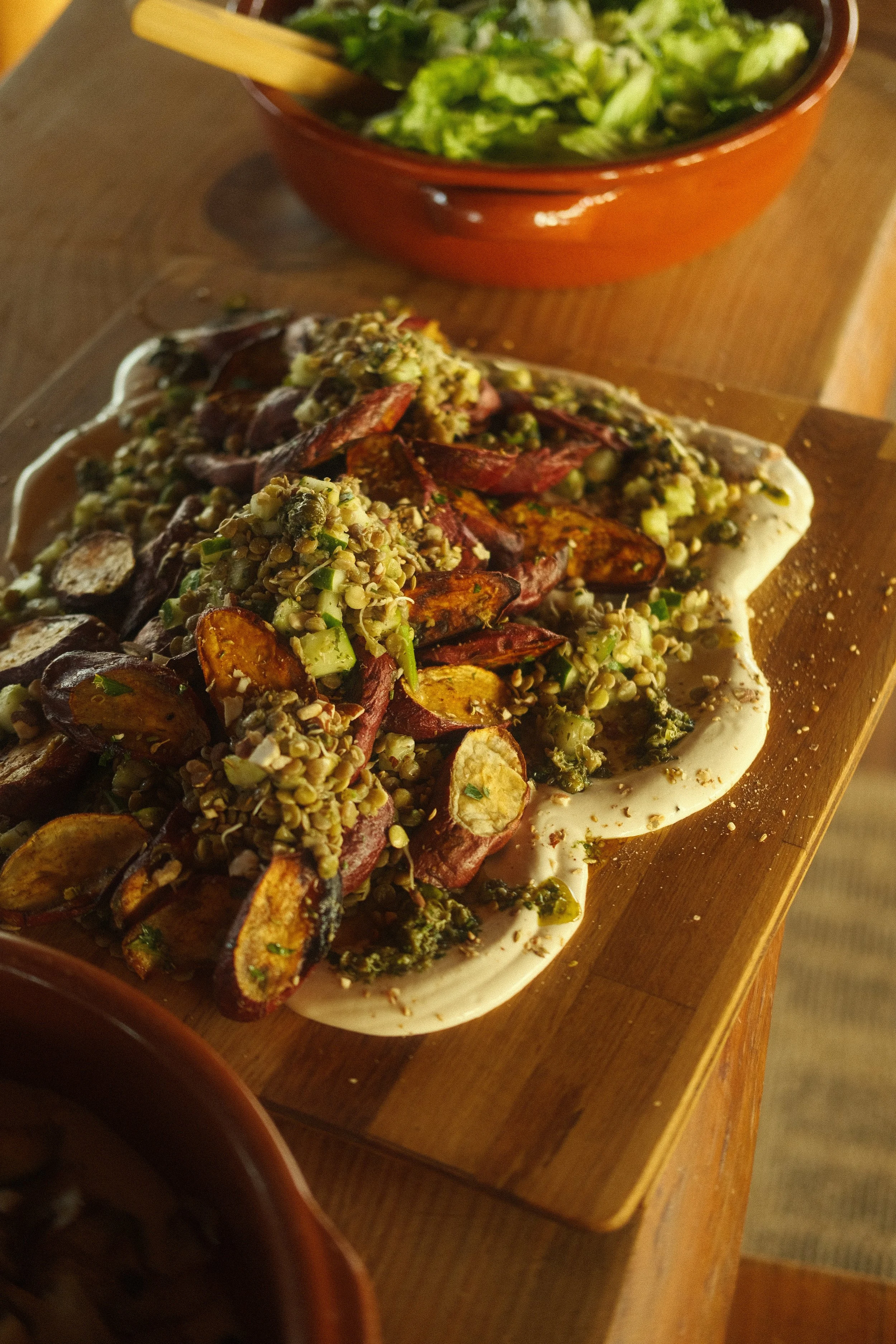 A plate of roasted mixed vegetables topped with green herbs, served on a white dish, with a bowl of chopped lettuce salad in the background.