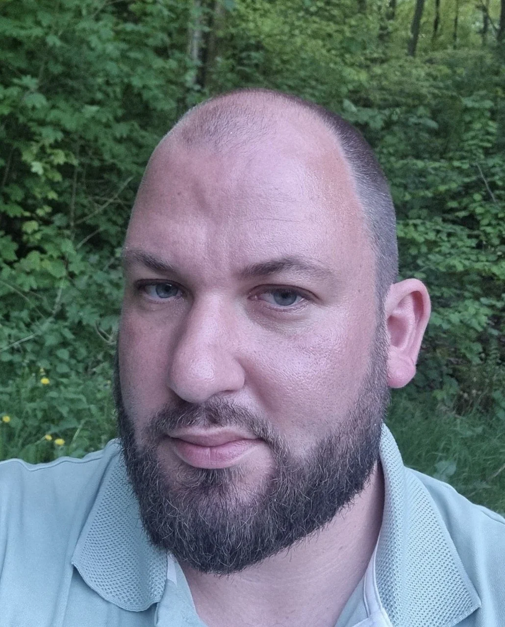 Close-up of a man with a beard and blue eyes, outdoors with green foliage in the background.