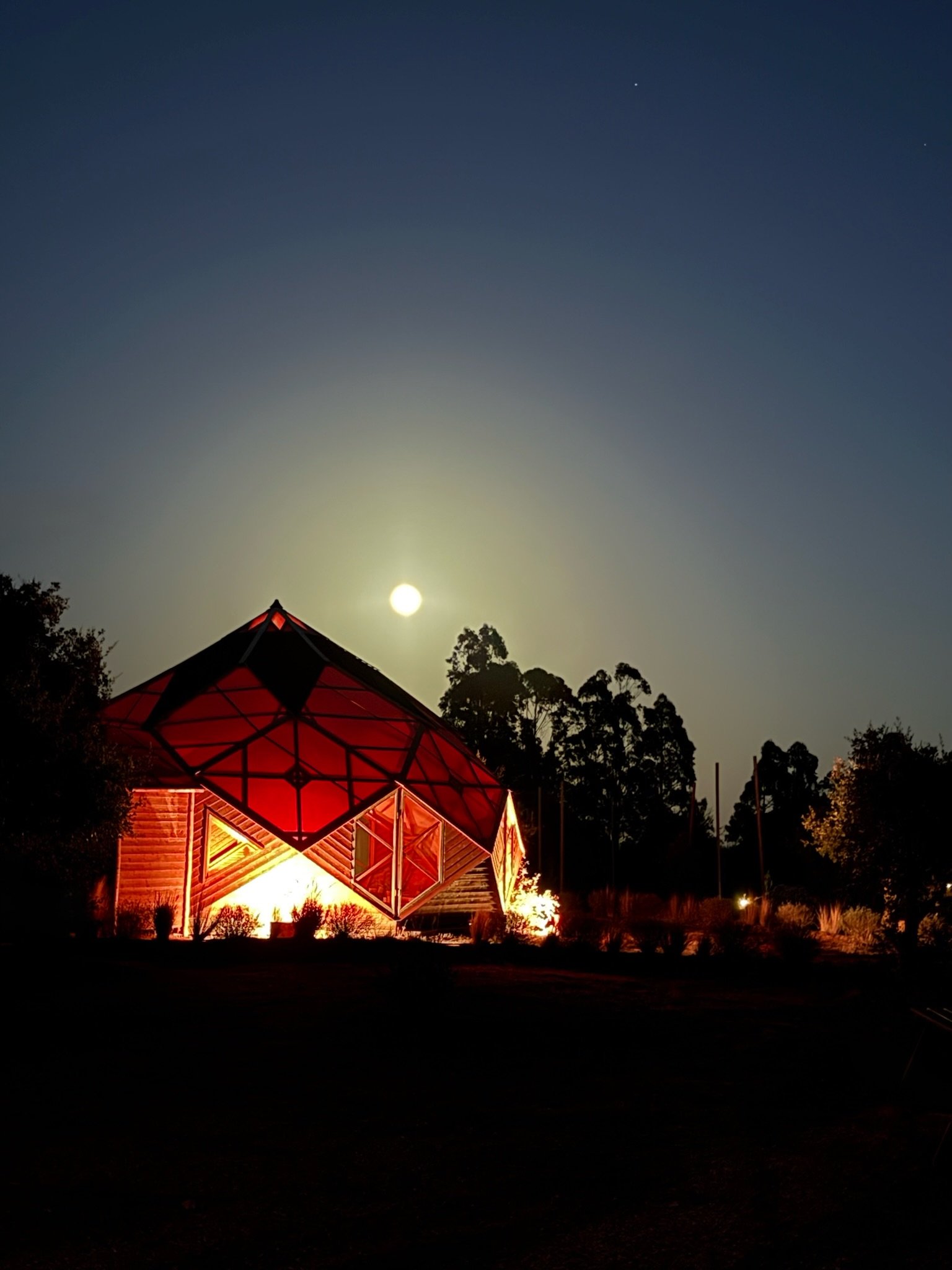 A geodesic dome structure illuminated with red lights, set against a nighttime sky with a bright full moon and silhouetted trees in the background.