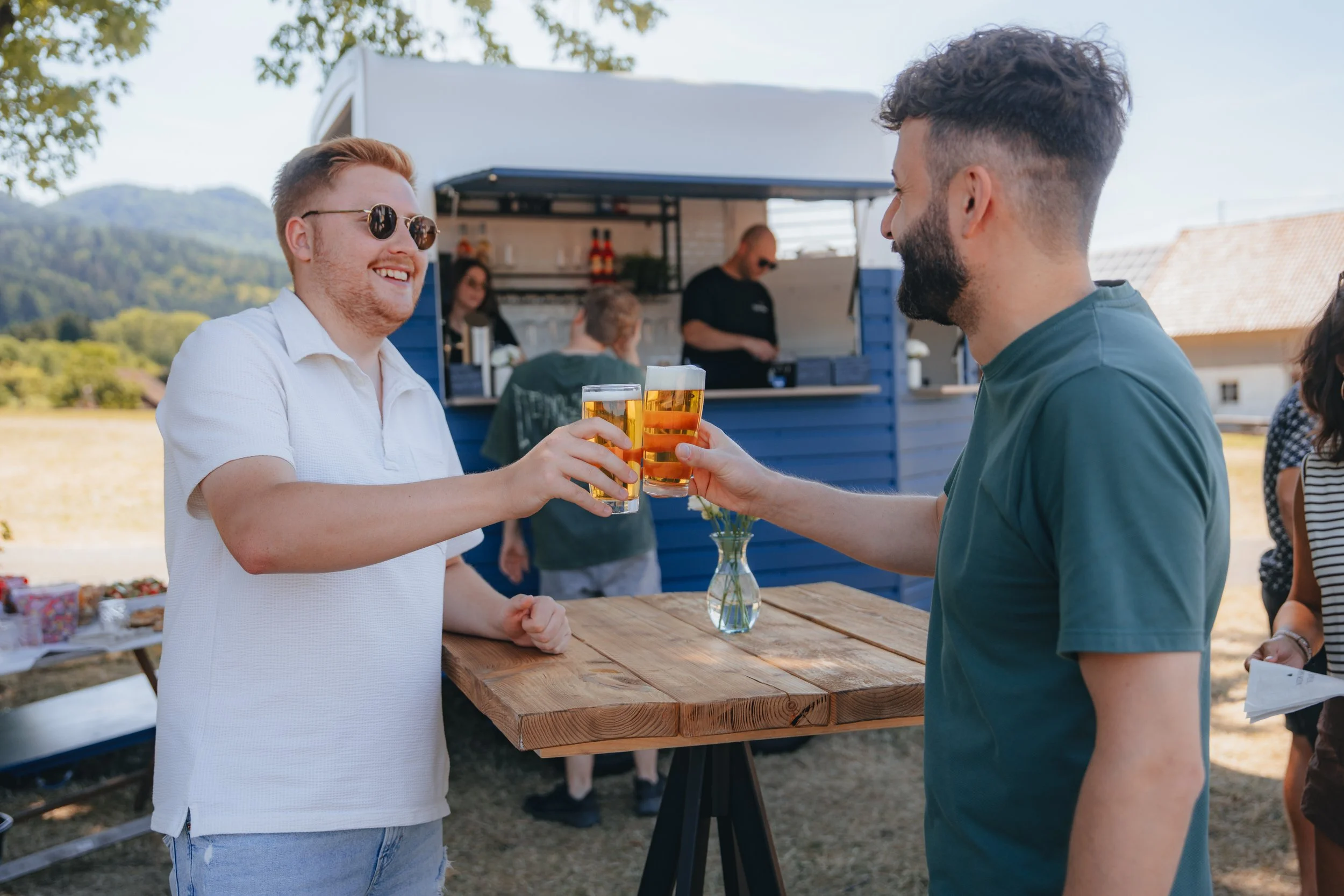 Bar mit verschiedenen Cocktails und Getränken, eine Person gießt einen Drink in ein Glas, im Hintergrund sind Menschen und große Fenster.