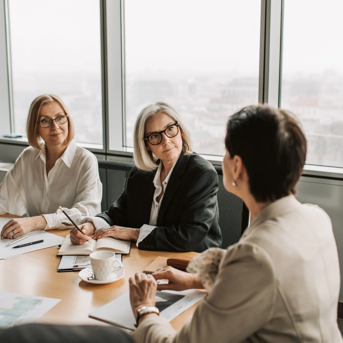 Three women in professional attire sitting at a conference table engaged in a discussion, with large windows and an urban cityscape in the background.