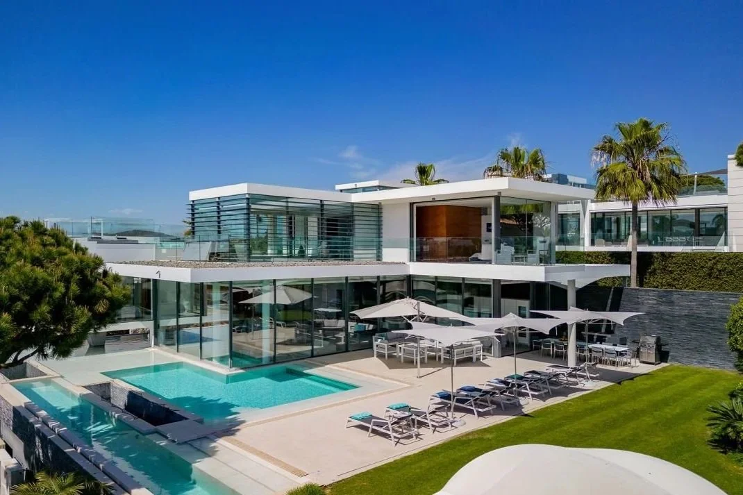 Modern house with large glass windows, outdoor pool, sun loungers, and palm trees under a blue sky.