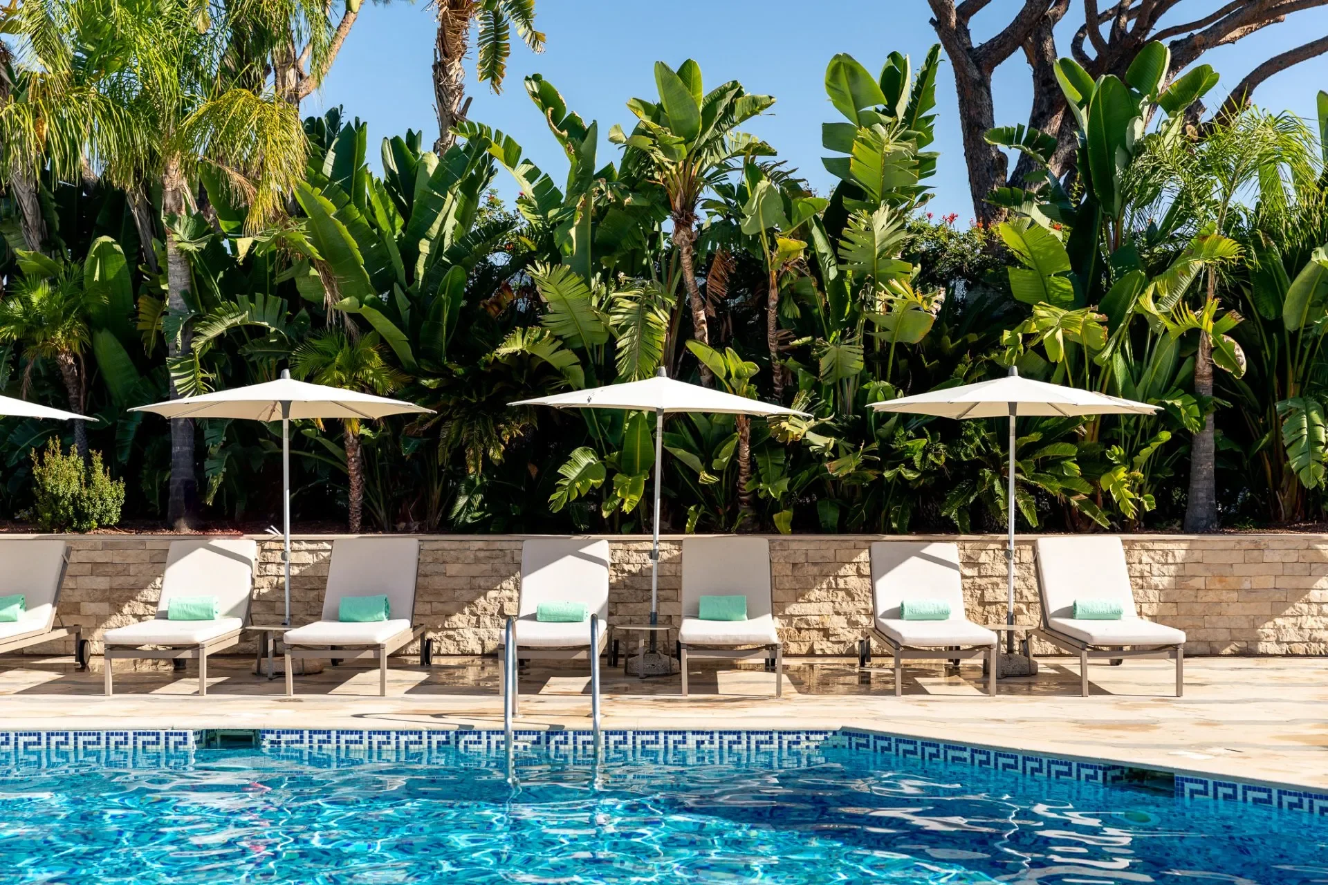 A woman walking on the edge of an infinity pool overlooking the ocean, wearing a large straw hat, pink top, and colorful, flowing cover-up.