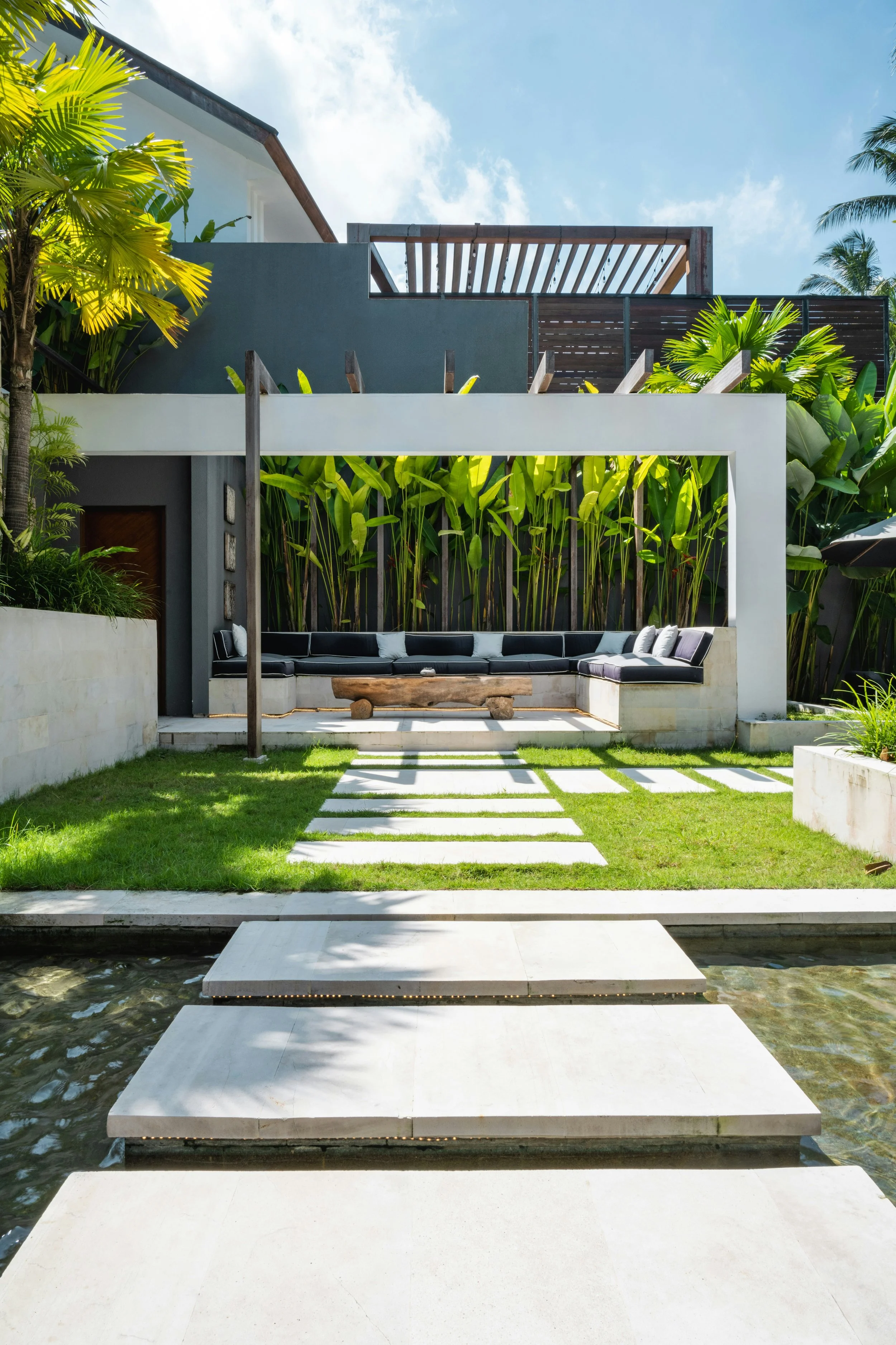 Modern backyard with a white pathway over water leading to a covered outdoor seating area with black cushions, surrounded by tropical plants and trees.