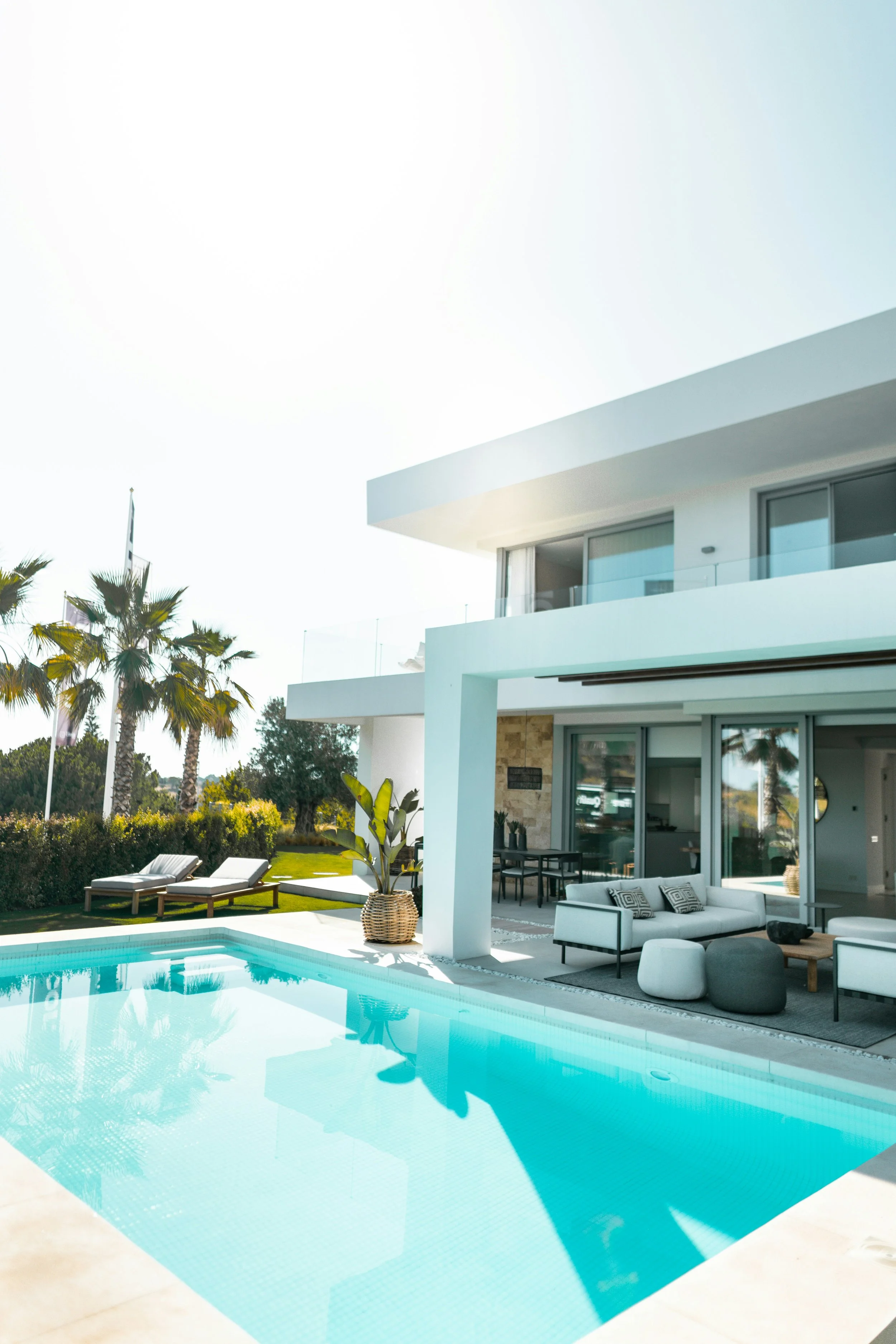 Luxury modern house with swimming pool, outdoor lounge area with white and gray furniture, tropical plants, and palm trees under clear sky.