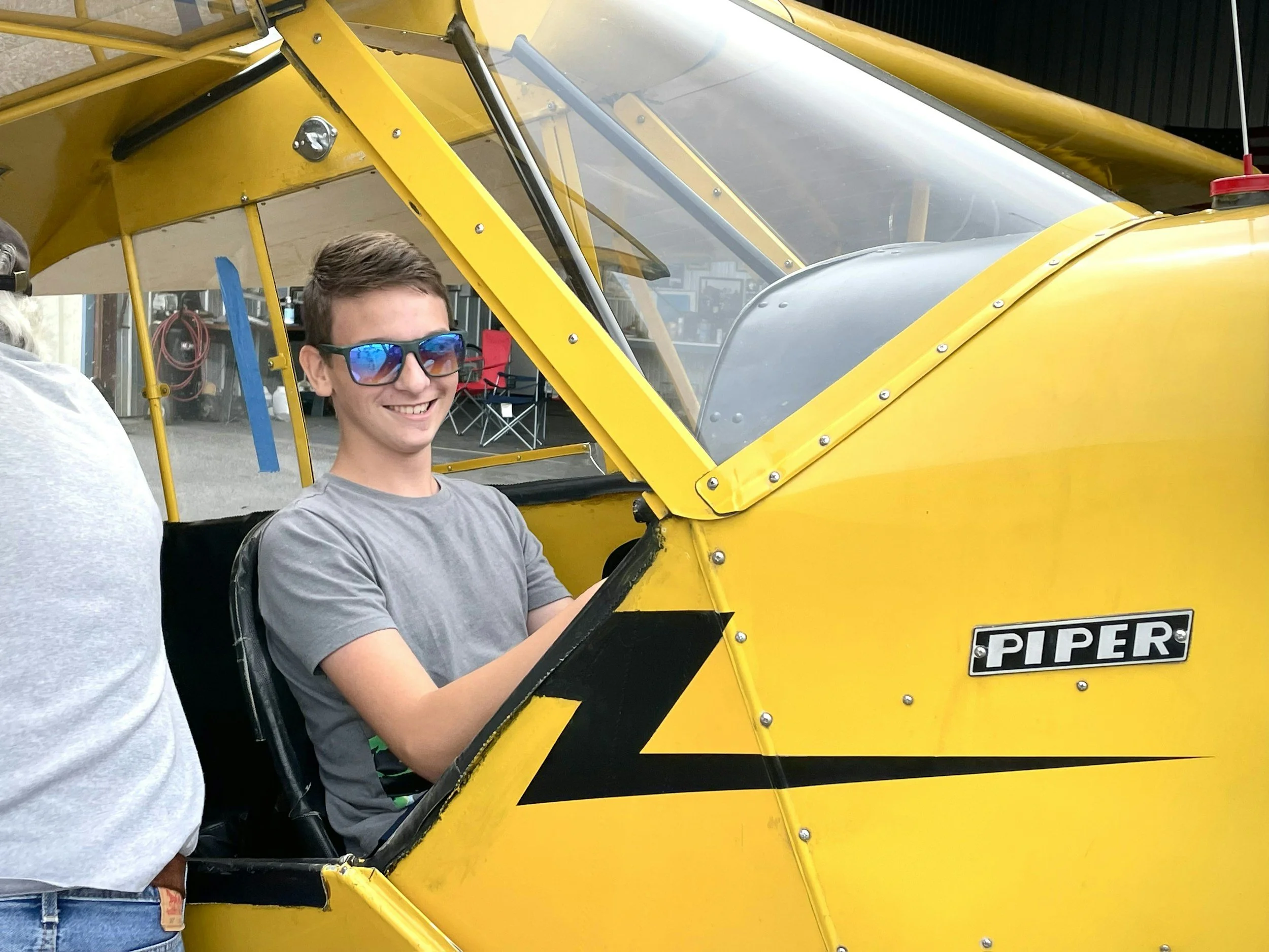 A young man wearing sunglasses sitting in the cockpit of a yellow Piper aircraft, smiling.