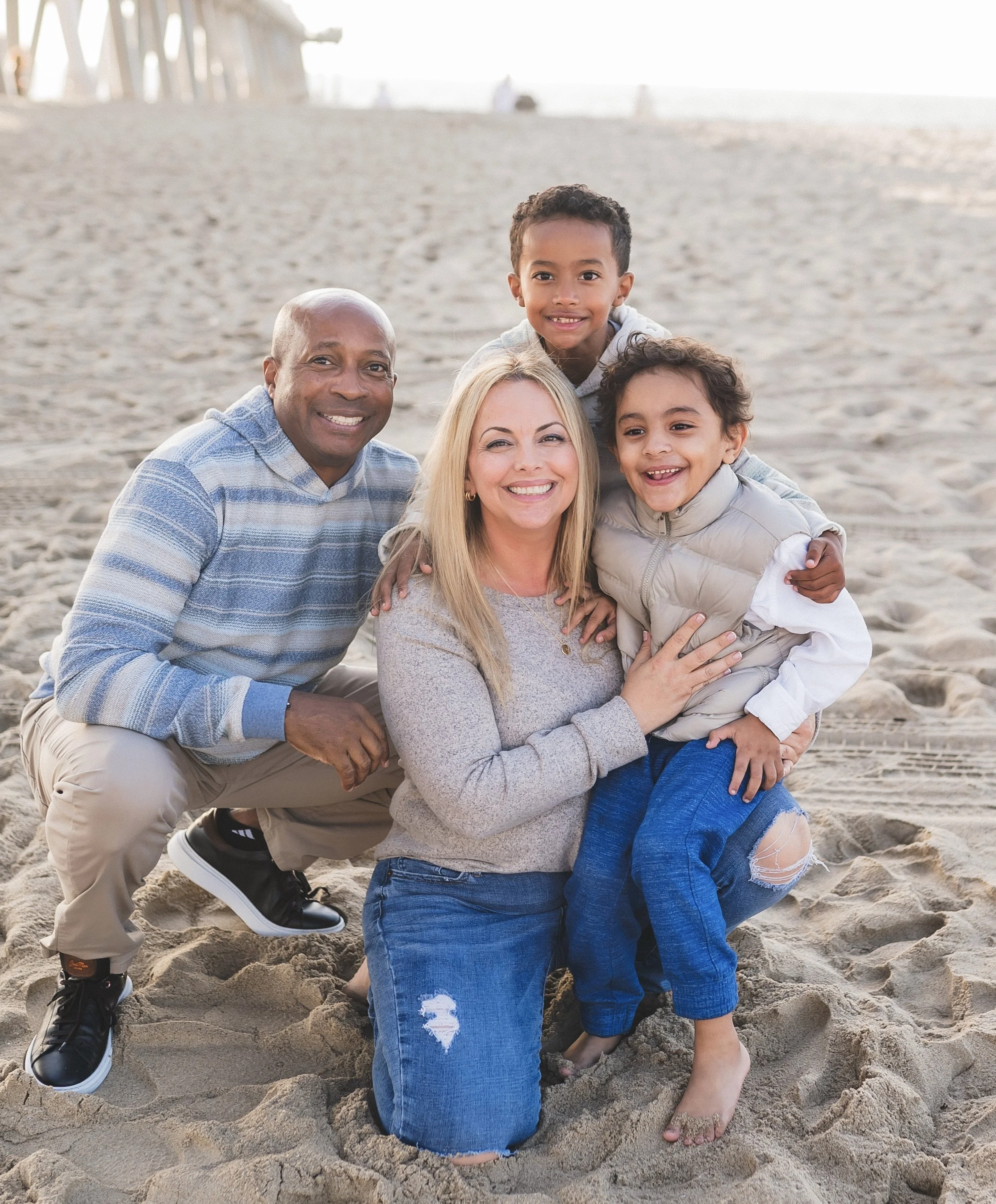 A family of four at the beach, smiling and posing for a photo. The mother is kneeling on the sand, with her children and father around her.