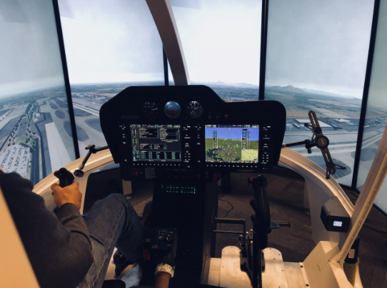 Inside a helicopter cockpit with two large windows showing the landscape outside, featuring controls, displays, and a person operating the aircraft.