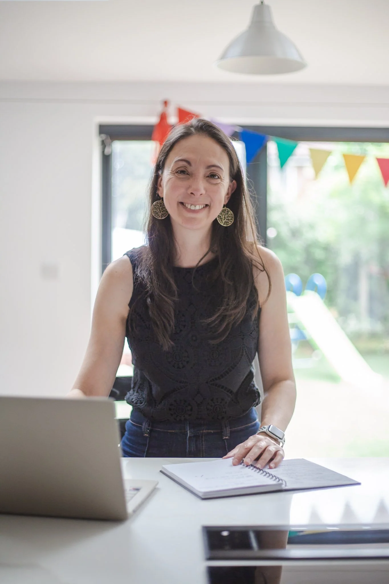 A woman with long dark hair smiling at a desk with laptops, notebooks, and a smartphone, in a room decorated for a celebration with colorful bunting and a slide in the background.