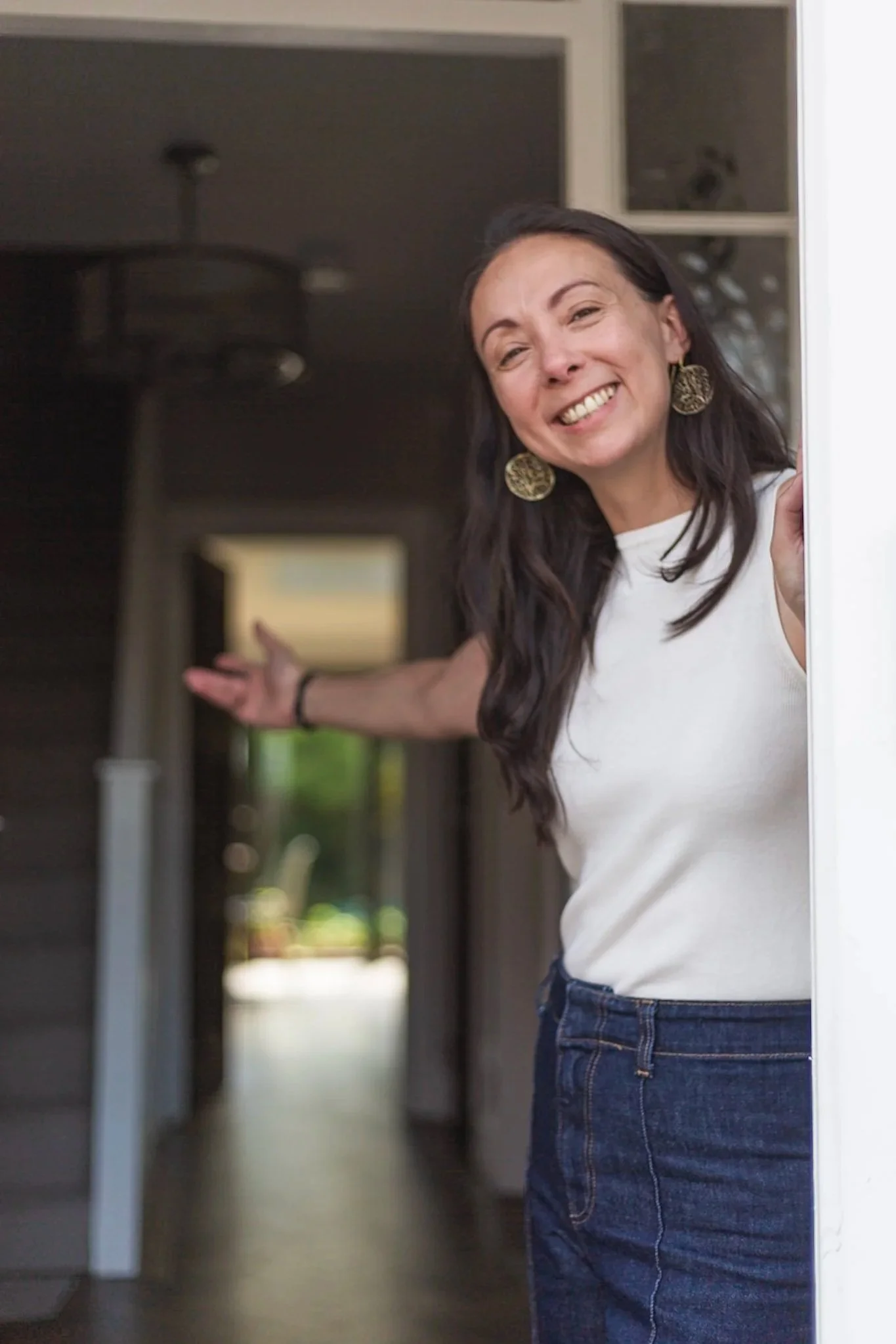 A woman smiling and waving from a doorway, wearing a white shirt, gold earrings, and jeans, with a blurred indoor background.