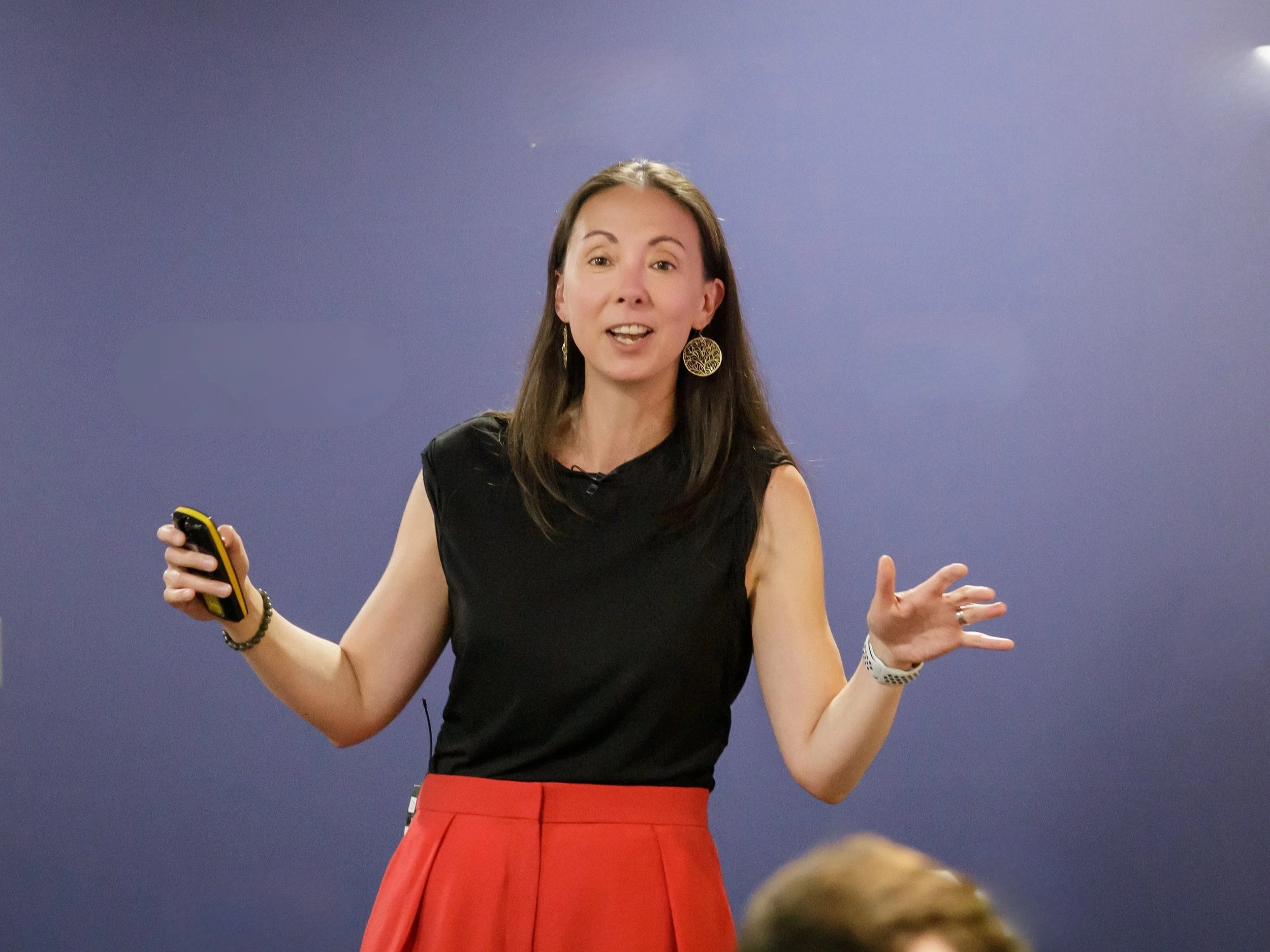 A woman with long brown hair wearing large earrings and a black top, speaking or presenting, gesturing with her hands, holding a remote or microphone, against a plain blue background.
