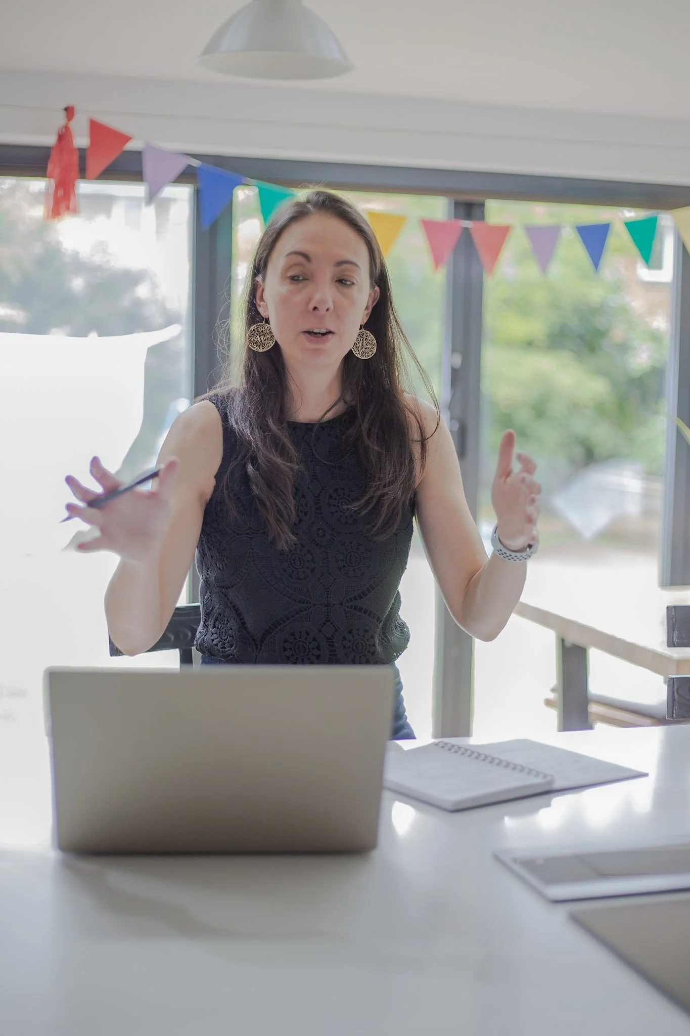 A woman with long brown hair wearing a black lace top and large earrings, gesturing as she speaks, sitting in a room decorated with colorful pennant banners.