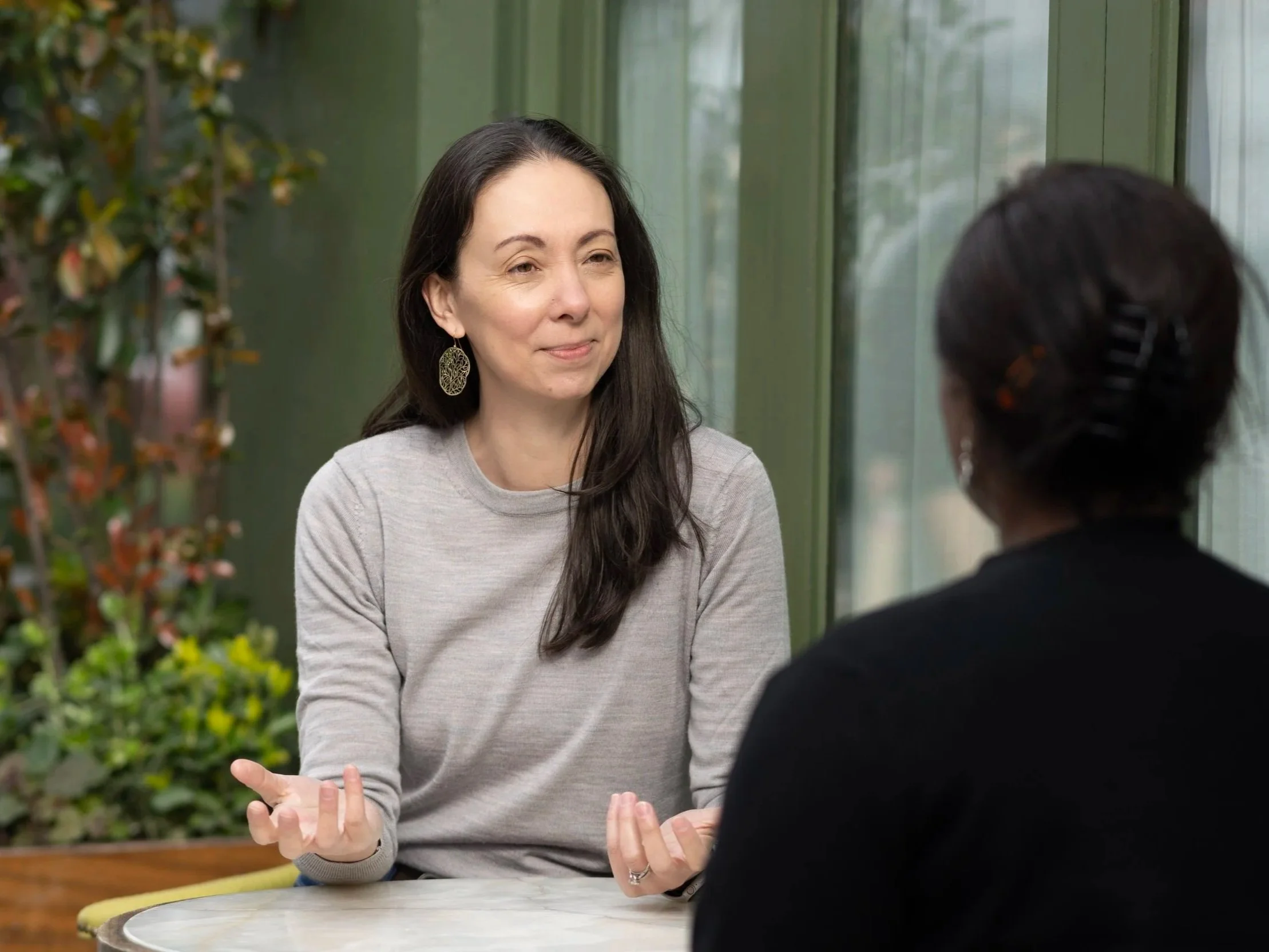 Two women are sitting at an outdoor table, engaged in conversation. One woman has long dark hair, is wearing earrings and a light-colored sweater, and is gesturing with her hands. The other woman has dark hair in a bun or covered with a hair clip, and wears a darker top. There are green plants and windows in the background.
