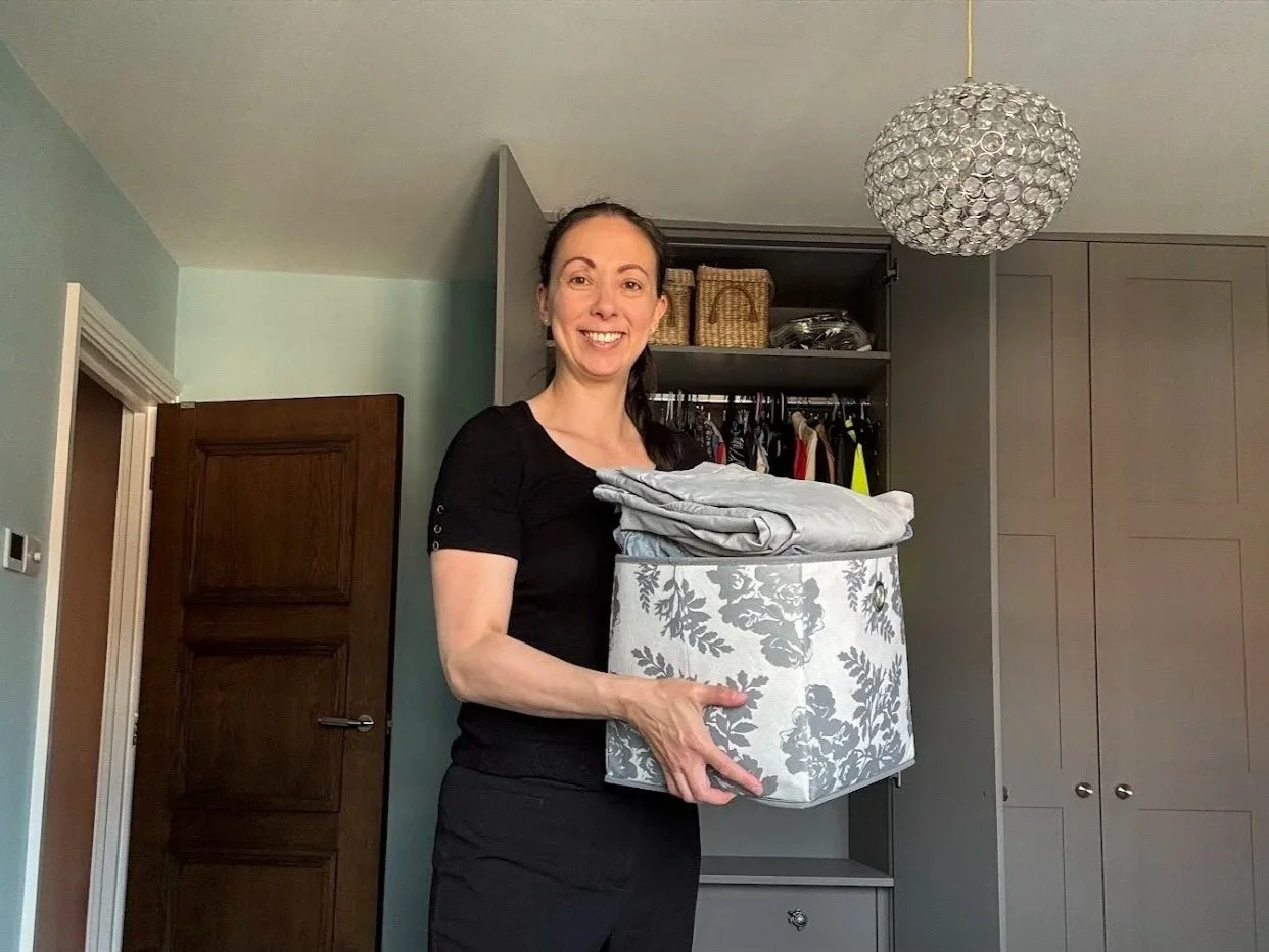 A woman smiling and holding a laundry basket with folded clothes inside in a bedroom