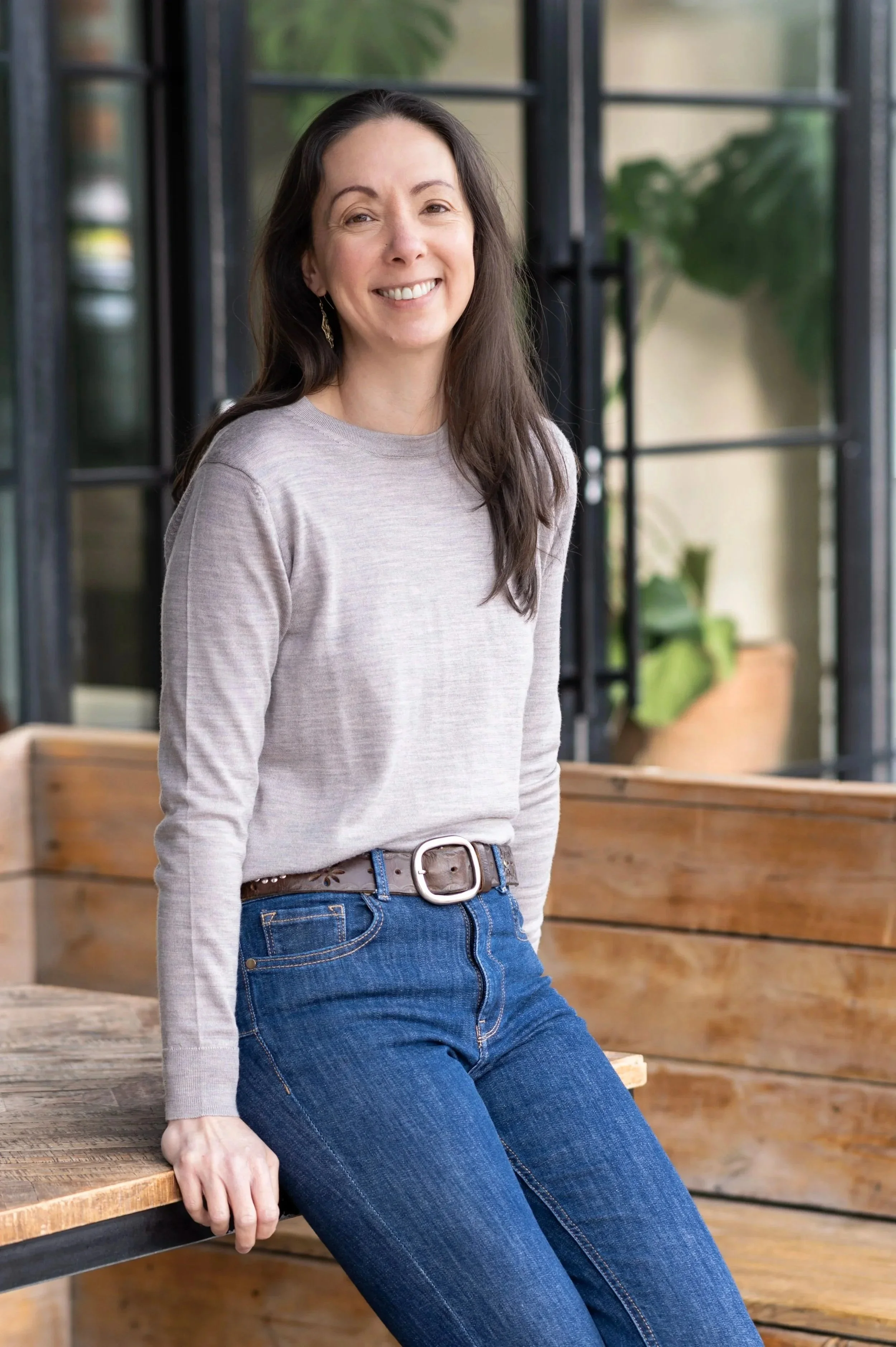 A woman with long dark hair, smiling, wearing a light gray long-sleeve shirt and blue jeans, sitting on a wooden bench in a well-lit indoor space with large windows and greenery outside.