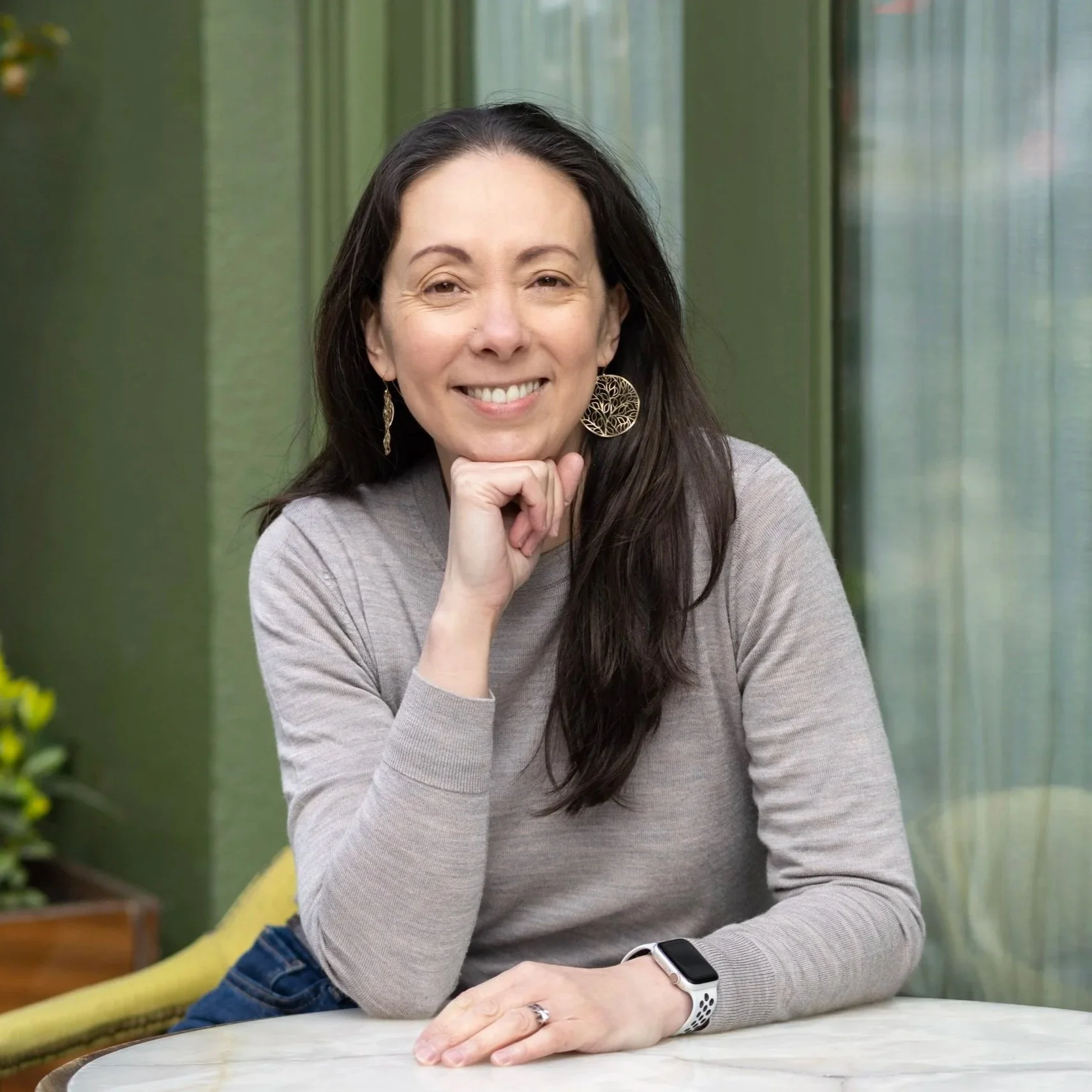 A woman with long dark hair, wearing a gray long sleeve shirt, large tree-shaped earrings, and a smartwatch, sits at a round table in a room with green walls, smiling at the camera, with her hand resting under her chin.