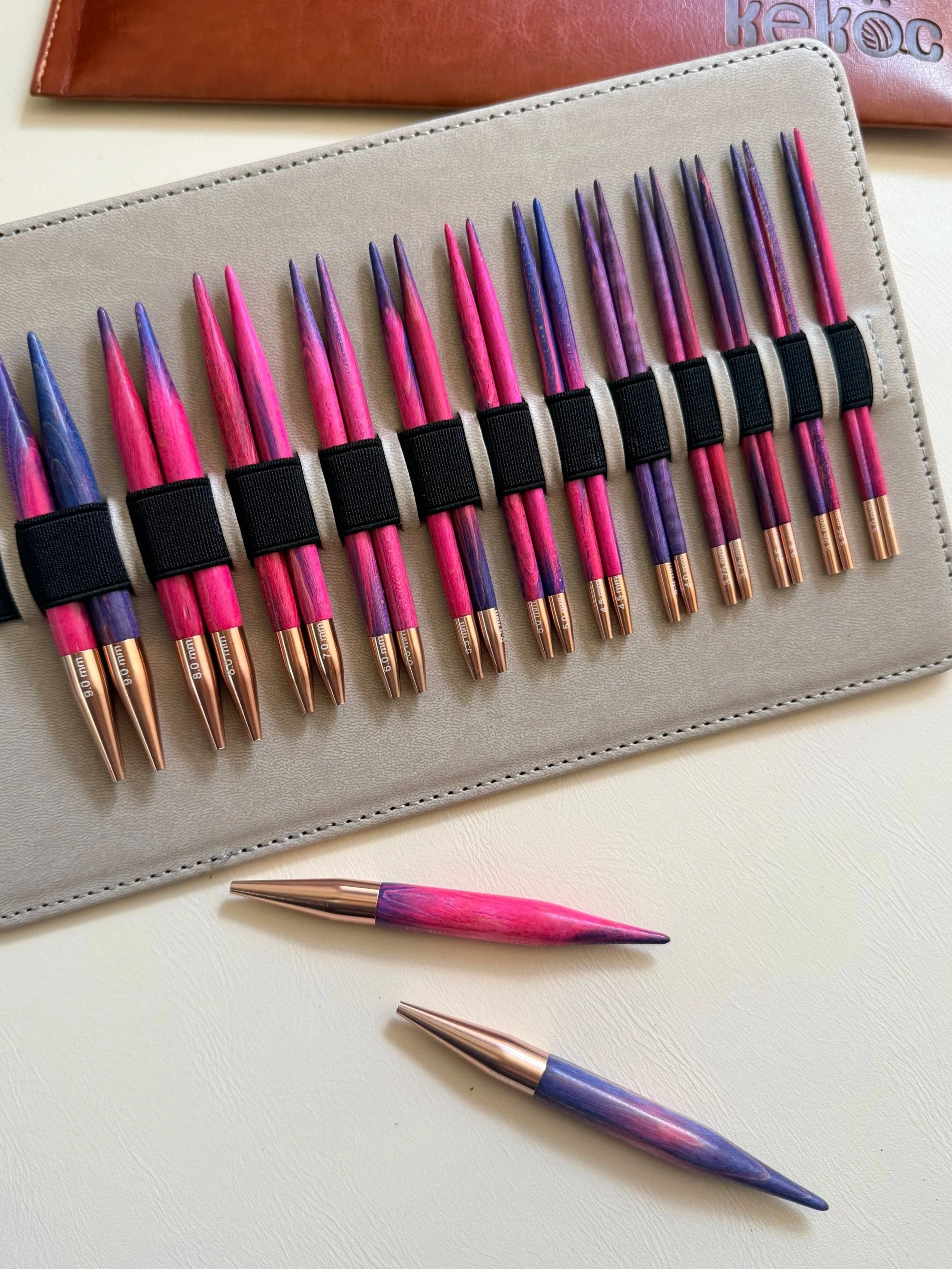 Set of knitting needles in pink, purple, and blue, arranged on a light-colored surface with a gray case holding some of the needles and a red book in the background.