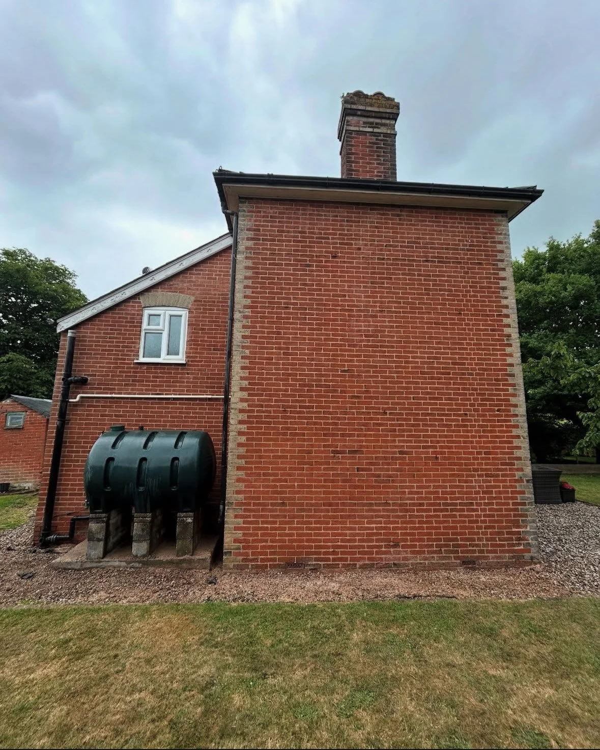 Backside of a brick house with a chimney, a small window, a black water tank, and a small lawn area under a cloudy sky.