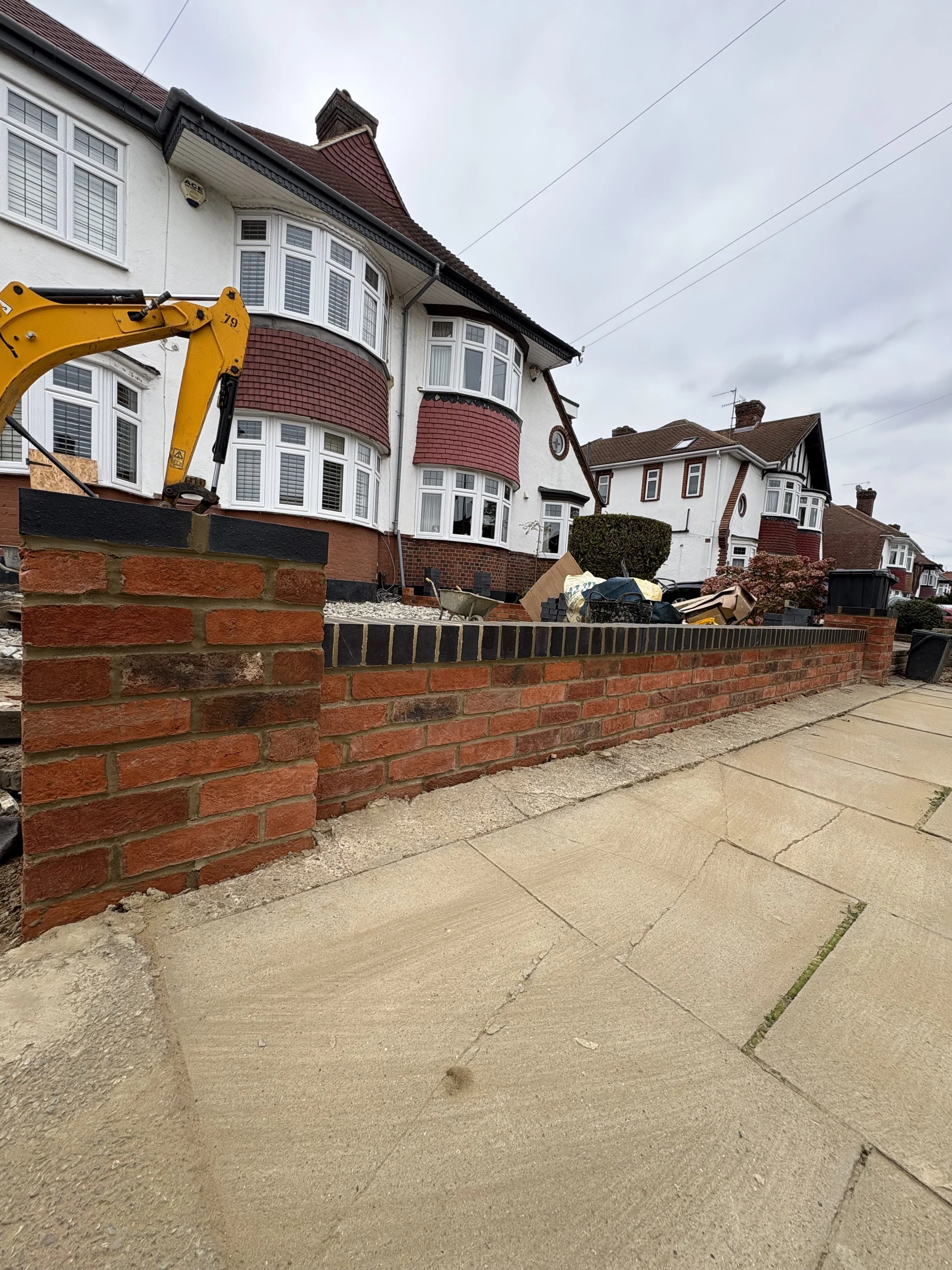 View of a residential street with a brick wall under construction in front of houses with bay windows, and construction equipment and materials on the sidewalk.